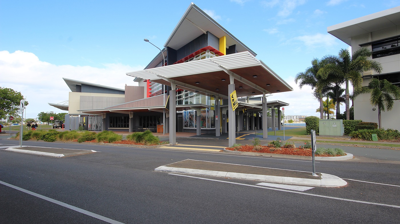 On the left, there is a curved road with a pavement and trees lining the side. On the right, there are modern buildings with large windows and some palm trees in front. In the middle, a building with a distinctive yellow vertical stripe and a slanted roof stands prominently. The background features more trees and parked cars. The foreground includes the road with white lane markings and a pedestrian crossing. On the left, there is a curved road with a pavement and trees lining the side. On the right, there are modern buildings with large windows and some palm trees in front. In the middle, a building with a distinctive yellow vertical stripe and a slanted roof stands prominently. The background features more trees and parked cars. The foreground includes the road with white lane markings and a pedestrian crossing.