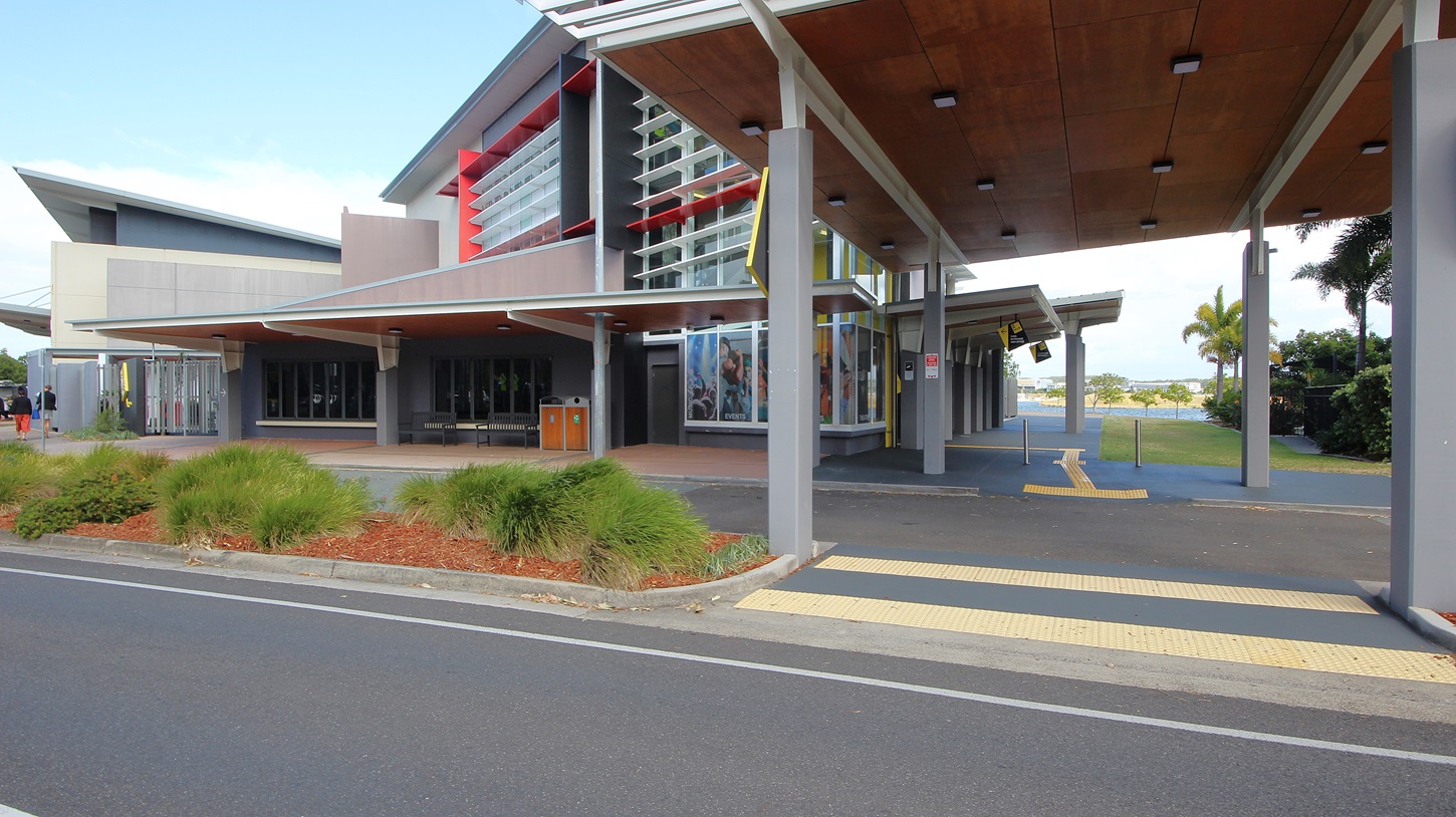 On the left, there is a modern building with large windows and a slanted roof, surrounded by landscaped areas with red mulch and shrubs. On the right, there is a covered walkway with a wooden ceiling and ceiling lights, leading to another building with palm trees nearby. In the middle, a prominent building with a yellow vertical stripe and large overhang extends towards the foreground. The foreground features a road with white lane markings and a pedestrian crossing with yellow tactile paving. On the left, there is a modern building with large windows and a slanted roof, surrounded by landscaped areas with red mulch and shrubs. On the right, there is a covered walkway with a wooden ceiling and ceiling lights, leading to another building with palm trees nearby. In the middle, a prominent building with a yellow vertical stripe and large overhang extends towards the foreground. The foreground features a road with white lane markings and a pedestrian crossing with yellow tactile paving.