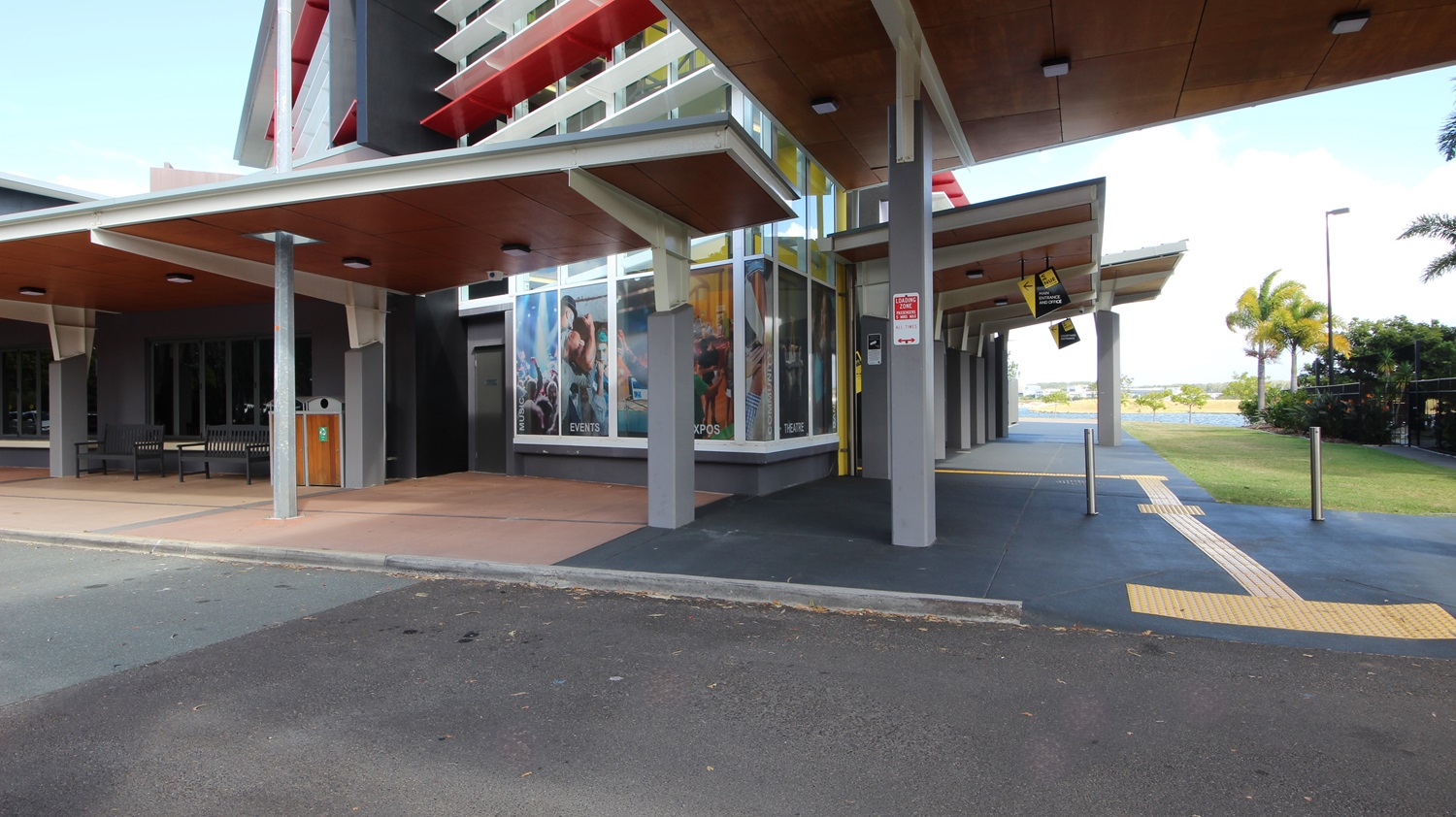 On the left, there is a building with large windows displaying posters and a wooden ceiling over a walkway. On the right, there is a covered area with a wooden ceiling and ceiling lights, leading to a pathway with yellow tactile paving. In the middle, a building with a yellow vertical stripe and red accents extends towards the foreground. The foreground features a paved area with a mix of dark and light grey flooring, and a small section of asphalt road. On the left, there is a building with large windows displaying posters and a wooden ceiling over a walkway. On the right, there is a covered area with a wooden ceiling and ceiling lights, leading to a pathway with yellow tactile paving. In the middle, a building with a yellow vertical stripe and red accents extends towards the foreground. The foreground features a paved area with a mix of dark and light grey flooring, and a small section of asphalt road.