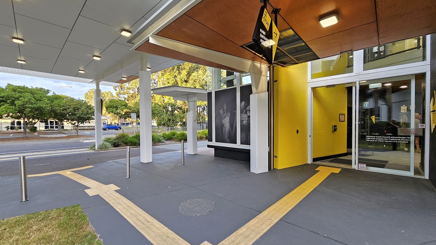 On the left, there is a covered walkway with a white ceiling and ceiling lights, leading to a building with large black-and-white posters. On the right, there is a glass entrance with a yellow wall and a door. In the middle, a yellow vertical stripe and a black overhang with a sign extend towards the foreground. The foreground features a dark grey floor with yellow tactile paving and a small patch of grass. The background includes trees and a road with parked cars. On the left, there is a covered walkway with a white ceiling and ceiling lights, leading to a building with large black-and-white posters. On the right, there is a glass entrance with a yellow wall and a door. In the middle, a yellow vertical stripe and a black overhang with a sign extend towards the foreground. The foreground features a dark grey floor with yellow tactile paving and a small patch of grass. The background includes trees and a road with parked cars.
