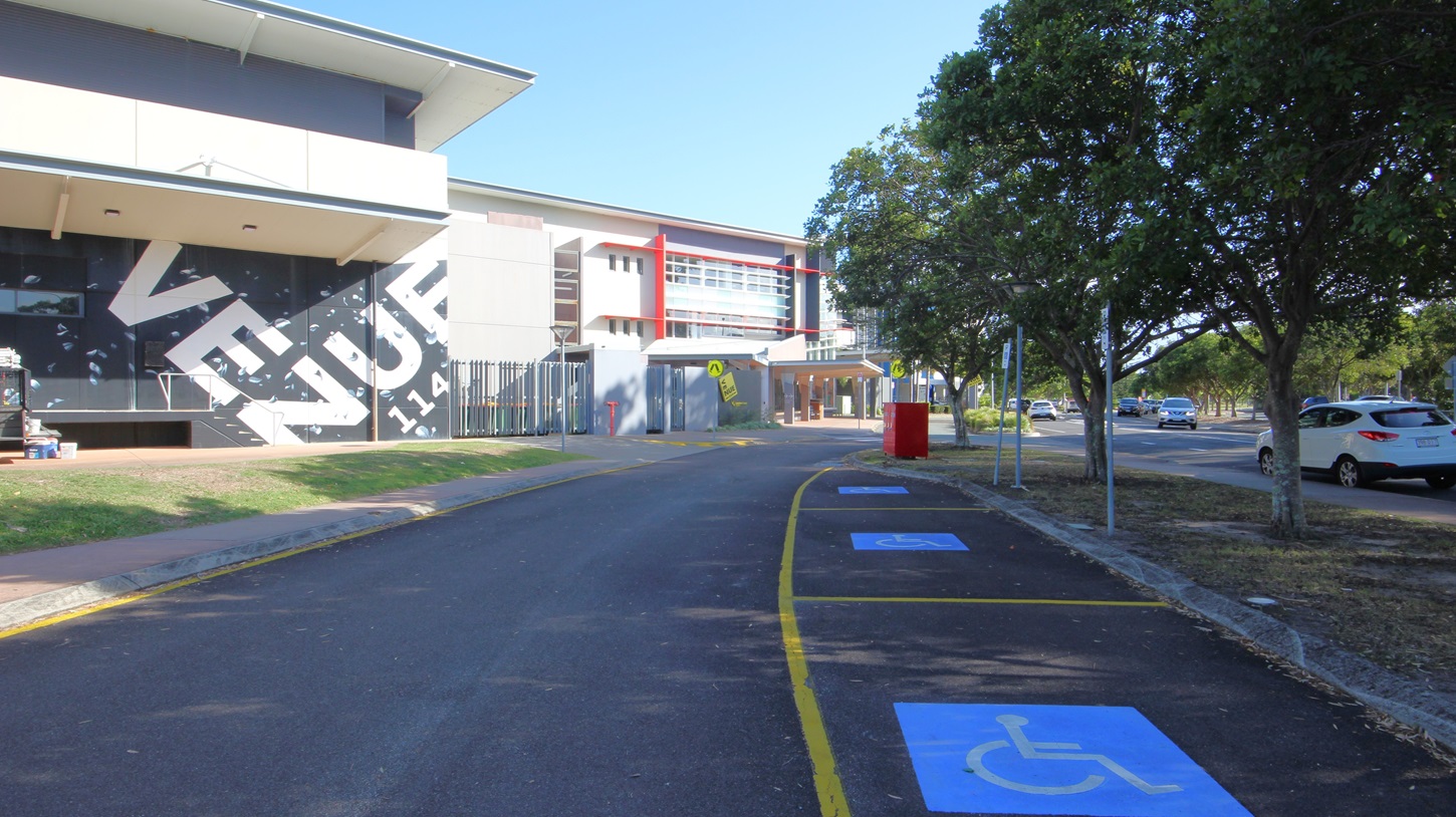 On the left, there is a building with a large sign reading "VENUE" in bold letters, featuring a slanted roof and a grassy area in front. On the right, there are several trees and parked cars along a road. In the middle, a building with red accents and multiple windows extends towards the background. The foreground features a paved road with yellow lines and blue disabled parking spaces marked with wheelchair symbols. On the left, there is a building with a large sign reading "VENUE" in bold letters, featuring a slanted roof and a grassy area in front. On the right, there are several trees and parked cars along a road. In the middle, a building with red accents and multiple windows extends towards the background. The foreground features a paved road with yellow lines and blue disabled parking spaces marked with wheelchair symbols.