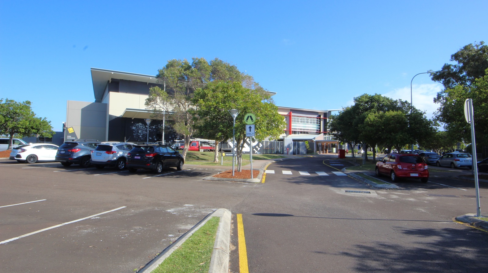 On the left, there are parked cars next to a building with a slanted roof and large windows. On the right, there are more parked cars and trees lining the area. In the middle, a building with red accents and multiple windows extends towards the background. The foreground features a paved road with white lane markings, a pedestrian crossing, and a small landscaped area with a tree and red mulch. On the left, there are parked cars next to a building with a slanted roof and large windows. On the right, there are more parked cars and trees lining the area. In the middle, a building with red accents and multiple windows extends towards the background. The foreground features a paved road with white lane markings, a pedestrian crossing, and a small landscaped area with a tree and red mulch.