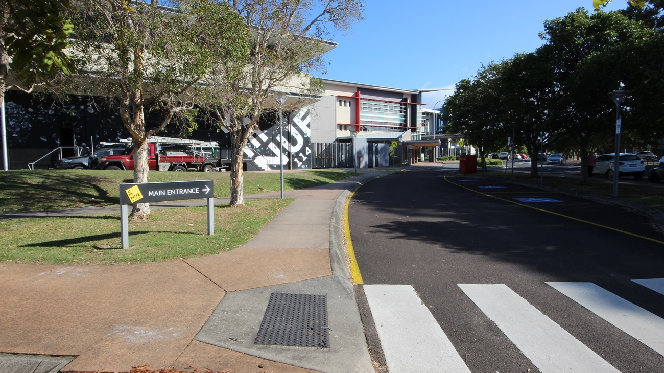 On the left, there is a building with a large mural, a tree, and a sign pointing to the main entrance. On the right, there are trees and parked cars along a road. In the middle, a building with red and grey accents and multiple windows extends towards the background. The foreground features a pedestrian crossing with white stripes, a paved walkway, and a section of asphalt road with yellow lines. On the left, there is a building with a large mural, a tree, and a sign pointing to the main entrance. On the right, there are trees and parked cars along a road. In the middle, a building with red and grey accents and multiple windows extends towards the background. The foreground features a pedestrian crossing with white stripes, a paved walkway, and a section of asphalt road with yellow lines.