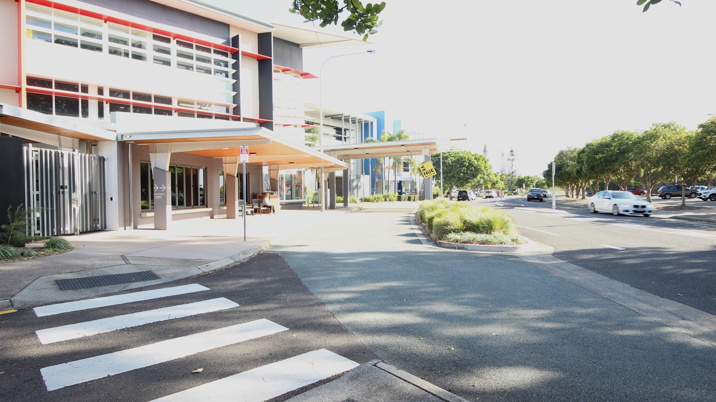 On the left, there is a modern building with red and grey accents, large windows, and a covered walkway with a beige ceiling. On the right, there are trees and parked cars along a road. In the middle, a continuation of the building extends towards the background, featuring more windows and a blue vertical stripe. The foreground includes a pedestrian crossing with white stripes, a paved walkway, and a section of asphalt road with a small landscaped area containing grass and shrubs. On the left, there is a modern building with red and grey accents, large windows, and a covered walkway with a beige ceiling. On the right, there are trees and parked cars along a road. In the middle, a continuation of the building extends towards the background, featuring more windows and a blue vertical stripe. The foreground includes a pedestrian crossing with white stripes, a paved walkway, and a section of asphalt road with a small landscaped area containing grass and shrubs.