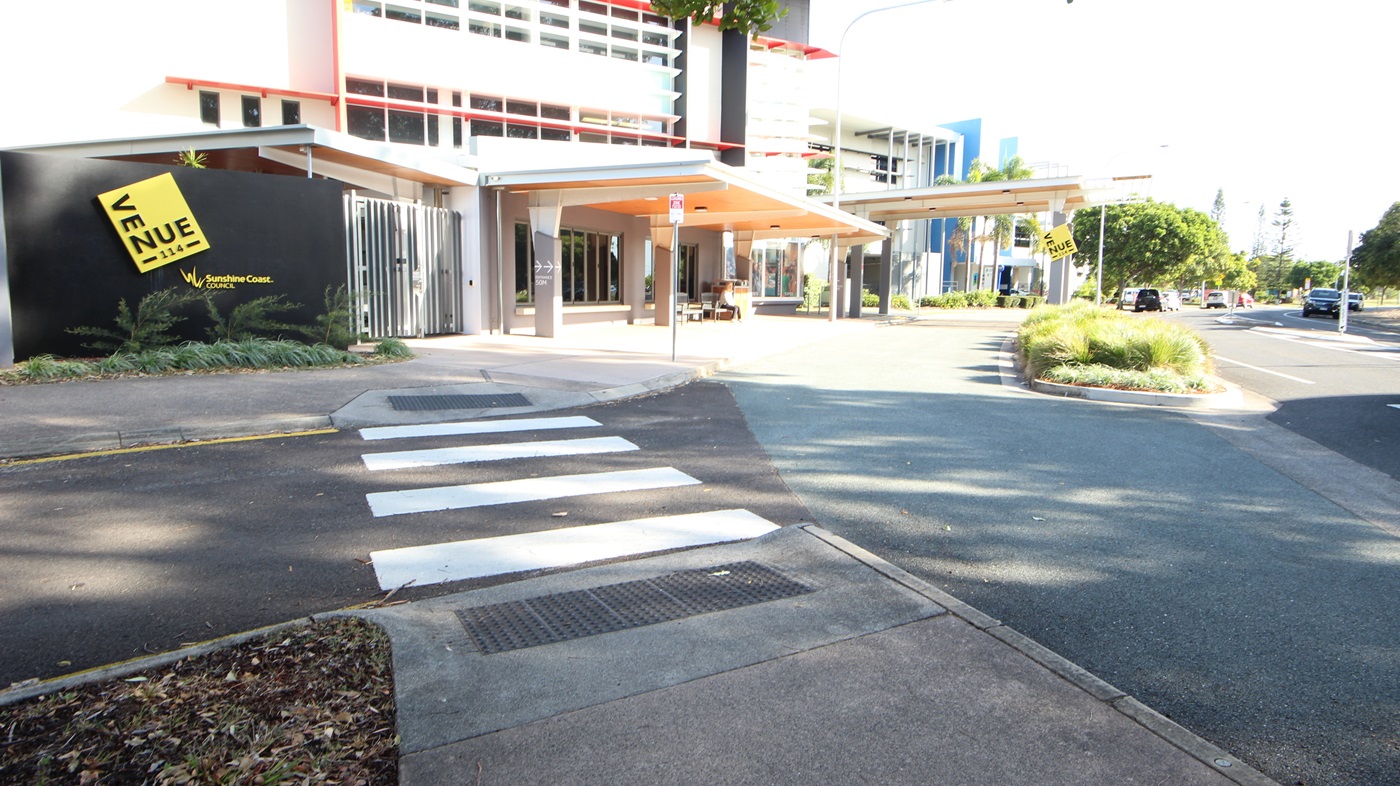 On the left, there is a building with a large yellow sign reading "VENUE" and a black wall with plants in front. On the right, there are trees and a road with parked cars. In the middle, a building with red and white accents and large windows extends towards the background, with a covered walkway featuring a beige ceiling. The foreground includes a pedestrian crossing with white stripes, a paved walkway, and a section of asphalt road with a small landscaped area containing grass and shrubs. On the left, there is a building with a large yellow sign reading "VENUE" and a black wall with plants in front. On the right, there are trees and a road with parked cars. In the middle, a building with red and white accents and large windows extends towards the background, with a covered walkway featuring a beige ceiling. The foreground includes a pedestrian crossing with white stripes, a paved walkway, and a section of asphalt road with a small landscaped area containing grass and shrubs.