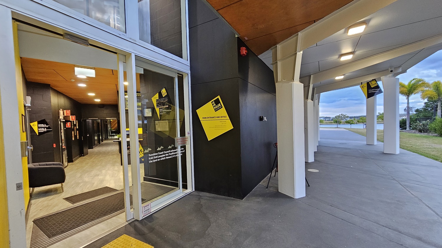 On the left, there is a glass entrance door with a yellow sign and a black sofa inside. The floor is light-coloured wood. On the right, there is a covered walkway with white pillars and a grey concrete floor. In the middle, a black wall with a yellow sign and a fire alarm. The background features a grassy area with palm trees and a body of water. The ceiling is wooden on the left and white on the right. On the left, there is a glass entrance door with a yellow sign and a black sofa inside. The floor is light-coloured wood. On the right, there is a covered walkway with white pillars and a grey concrete floor. In the middle, a black wall with a yellow sign and a fire alarm. The background features a grassy area with palm trees and a body of water. The ceiling is wooden on the left and white on the right.
