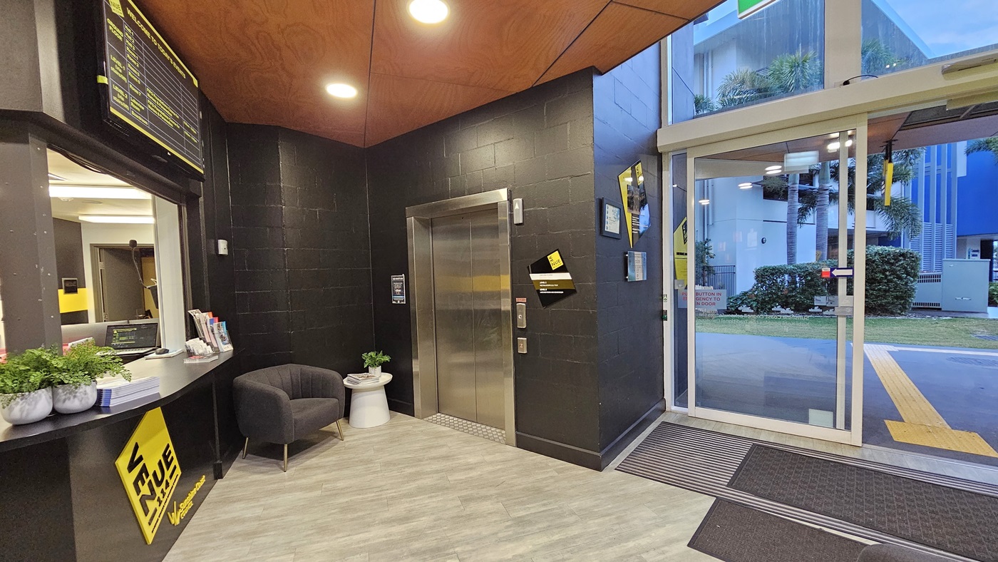 On the left, there is a reception desk with a yellow sign, a black chair, and a small white table with plants. On the right, there is a glass entrance door with a yellow sign and a view of a grassy area and buildings outside. In the middle, there is a lift with a yellow stripe and a black wall. The floor is light grey tile, and the ceiling is wooden with recessed lights. On the left, there is a reception desk with a yellow sign, a black chair, and a small white table with plants. On the right, there is a glass entrance door with a yellow sign and a view of a grassy area and buildings outside. In the middle, there is a lift with a yellow stripe and a black wall. The floor is light grey tile, and the ceiling is wooden with recessed lights.