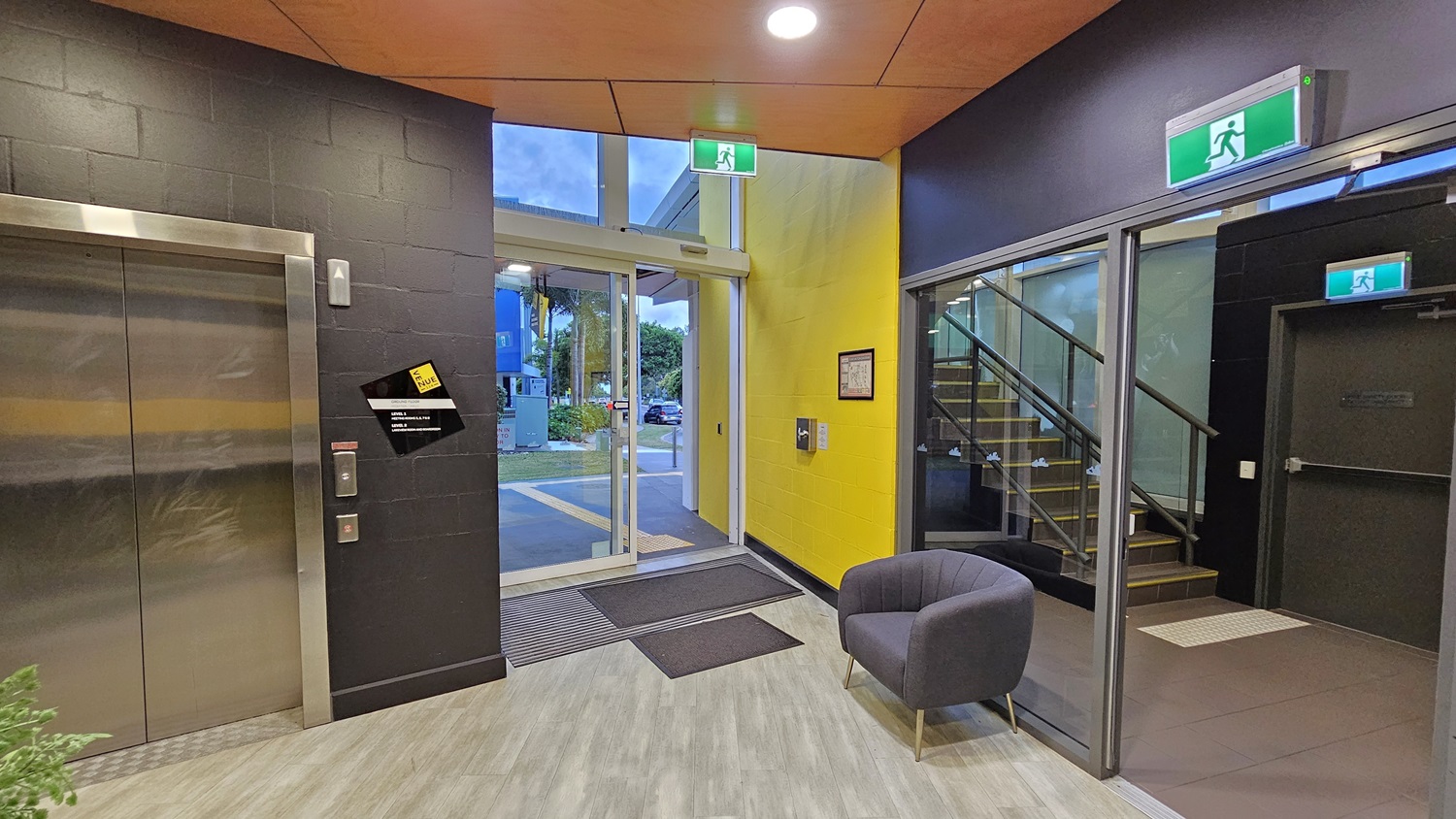 On the left, there is a lift with a black wall and a yellow stripe. On the right, there is a glass door with a green exit sign above it, leading to a staircase with a black railing. In the middle, there is a glass entrance door with a view of a car park and greenery outside. The floor is light grey wood-effect tile, and the ceiling is orange with recessed lights. A grey armchair is placed near the entrance. On the left, there is a lift with a black wall and a yellow stripe. On the right, there is a glass door with a green exit sign above it, leading to a staircase with a black railing. In the middle, there is a glass entrance door with a view of a car park and greenery outside. The floor is light grey wood-effect tile, and the ceiling is orange with recessed lights. A grey armchair is placed near the entrance.