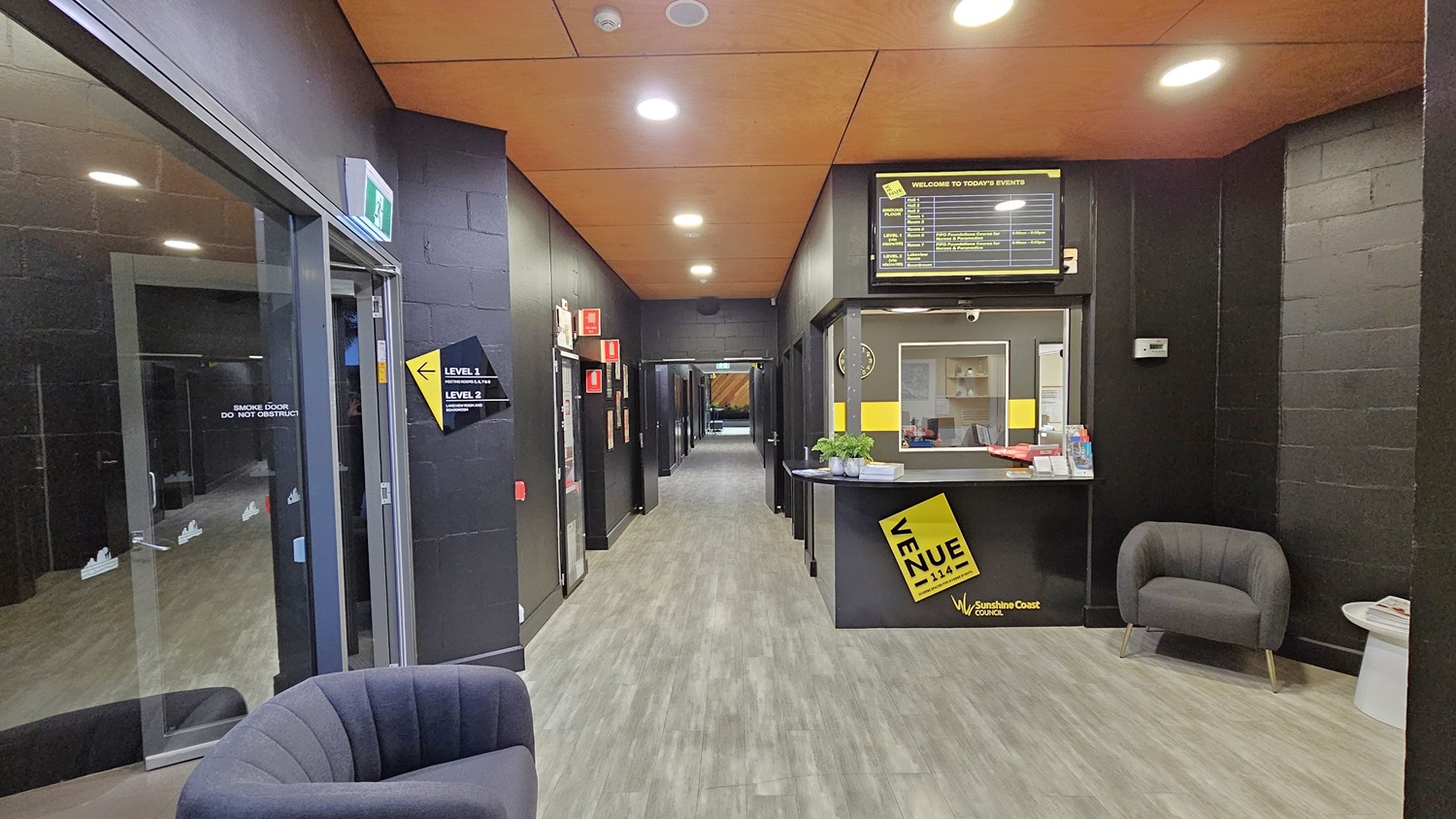 On the left, there is a glass door with a green exit sign above it, leading to a corridor with black walls. On the right, there is a reception desk with a yellow sign, a computer, and a small counter with brochures. In the middle, there is a hallway with black walls and several doors. The floor is grey wood-effect tile, and the ceiling is orange with recessed lights. Two grey armchairs are placed near the entrance. On the left, there is a glass door with a green exit sign above it, leading to a corridor with black walls. On the right, there is a reception desk with a yellow sign, a computer, and a small counter with brochures. In the middle, there is a hallway with black walls and several doors. The floor is grey wood-effect tile, and the ceiling is orange with recessed lights. Two grey armchairs are placed near the entrance.