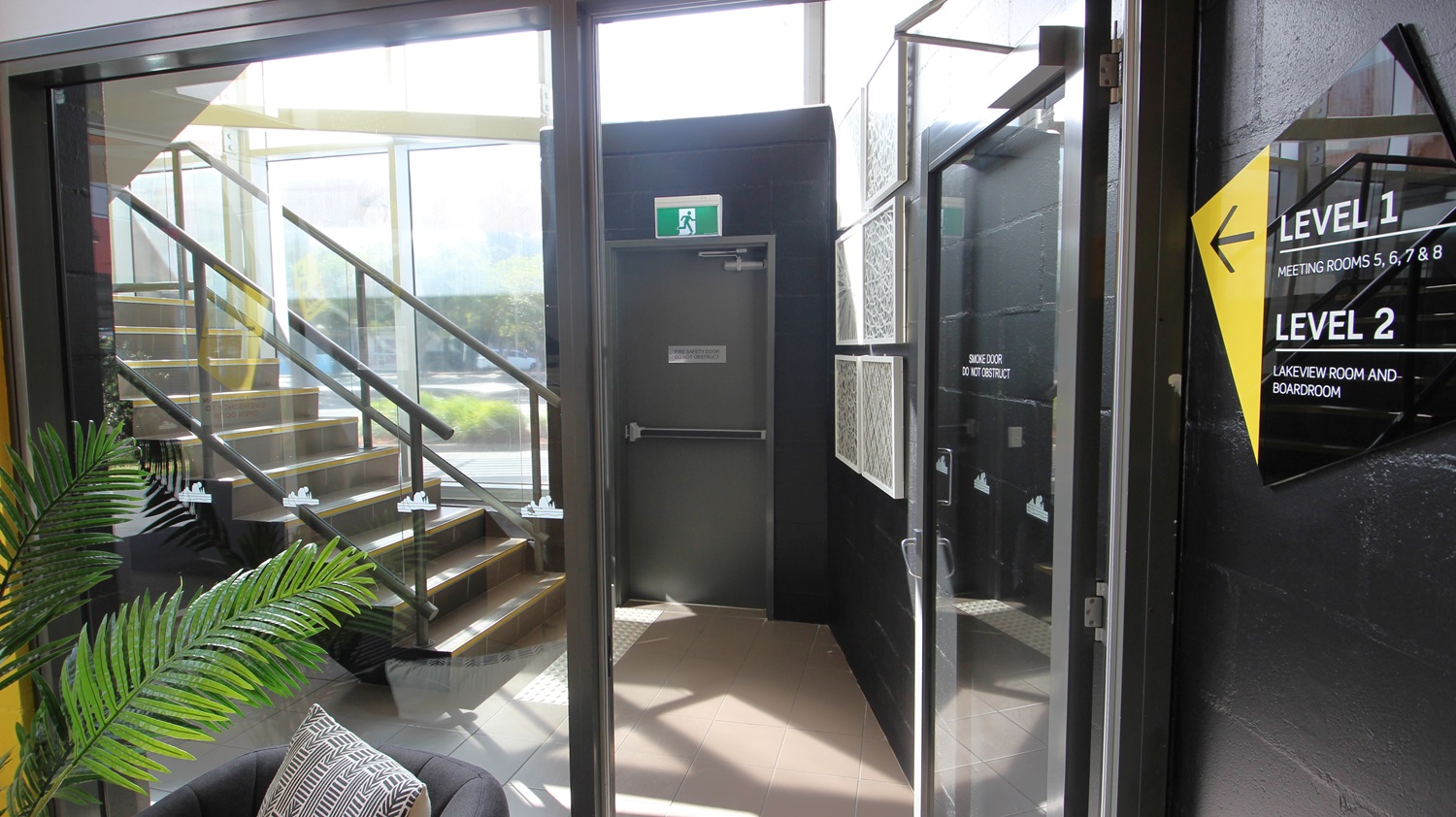 On the left, there is a staircase with black railings and wooden steps, leading to an upper level. On the right, there is a black wall with a yellow and black sign indicating "Level 1" and "Level 2". In the middle, there is a grey door with a green exit sign above it. The floor is light brown tile, and the ceiling is glass, allowing natural light to enter. A black armchair with a patterned cushion is in the foreground near a potted plant. On the left, there is a staircase with black railings and wooden steps, leading to an upper level. On the right, there is a black wall with a yellow and black sign indicating "Level 1" and "Level 2". In the middle, there is a grey door with a green exit sign above it. The floor is light brown tile, and the ceiling is glass, allowing natural light to enter. A black armchair with a patterned cushion is in the foreground near a potted plant.