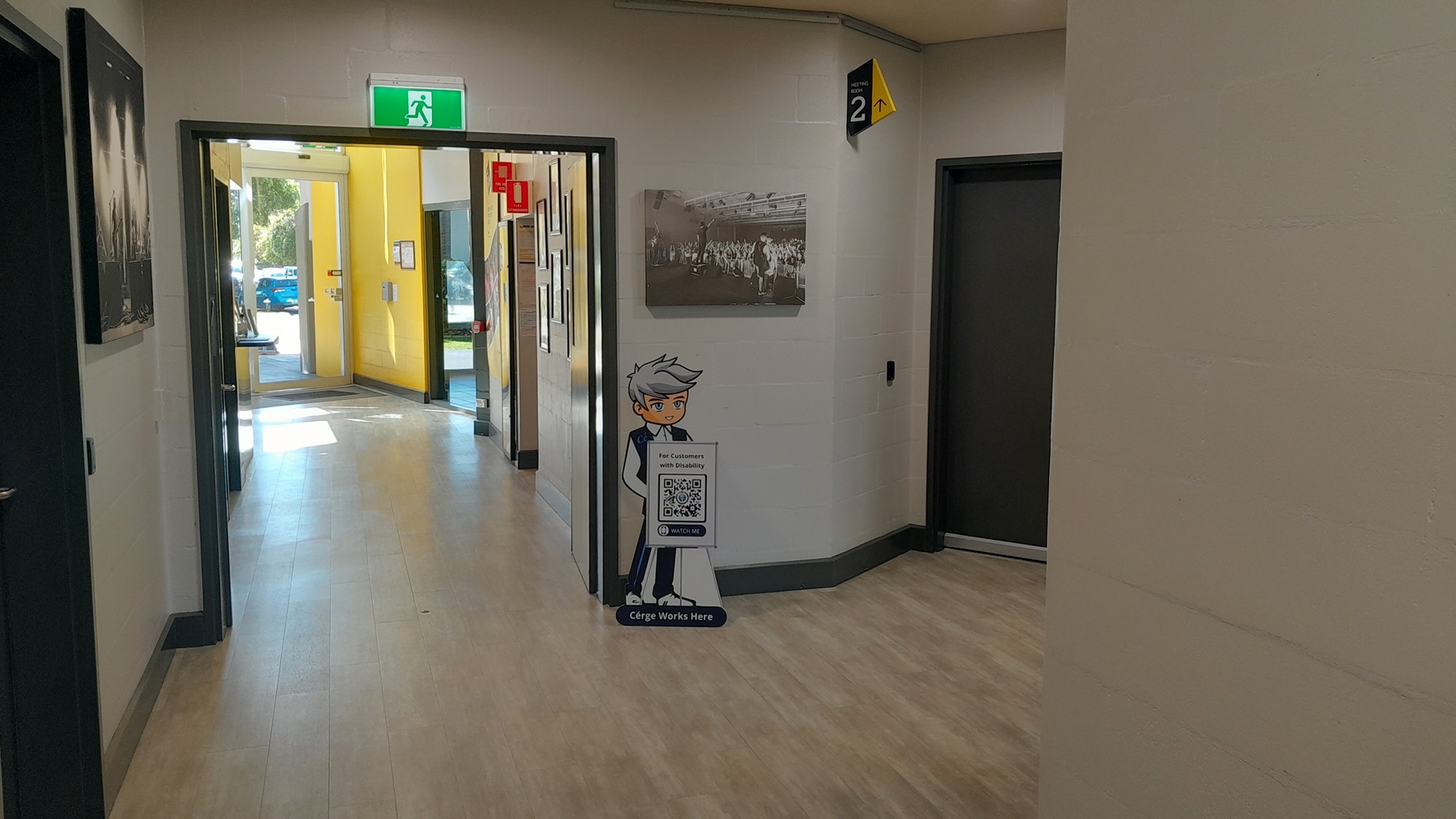 On the left, there is a black and white photograph on the wall and a corridor leading to a room with yellow walls. On the right, there is a black door with a yellow and black "Level 2" sign above it. In the middle, there is a green exit sign above a glass door leading outside, with a standee of a cartoon character holding a QR code. The floor is light wood-effect tile, and the ceiling is white with recessed lights. On the left, there is a black and white photograph on the wall and a corridor leading to a room with yellow walls. On the right, there is a black door with a yellow and black "Level 2" sign above it. In the middle, there is a green exit sign above a glass door leading outside, with a standee of a cartoon character holding a QR code. The floor is light wood-effect tile, and the ceiling is white with recessed lights.