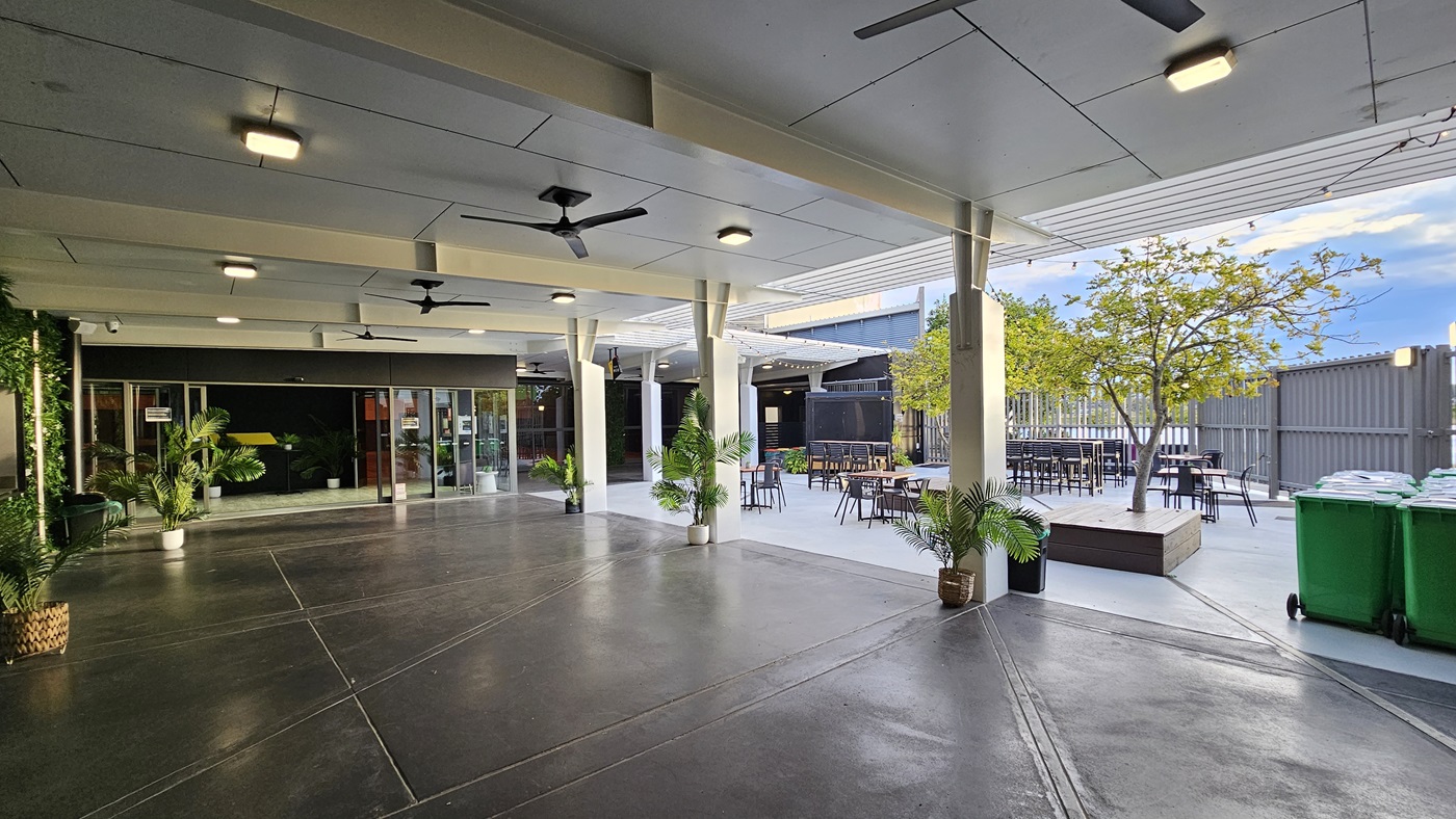 On the left, there is a glass entrance with potted plants and a black wall. On the right, there is an outdoor seating area with tables, chairs, and a tree in a planter box. In the middle, there is an open space with a concrete floor and several ceiling fans. The background features a grey fence and additional seating. The ceiling is white with recessed lights and fans. Green bins are placed near the fence on the right. On the left, there is a glass entrance with potted plants and a black wall. On the right, there is an outdoor seating area with tables, chairs, and a tree in a planter box. In the middle, there is an open space with a concrete floor and several ceiling fans. The background features a grey fence and additional seating. The ceiling is white with recessed lights and fans. Green bins are placed near the fence on the right.