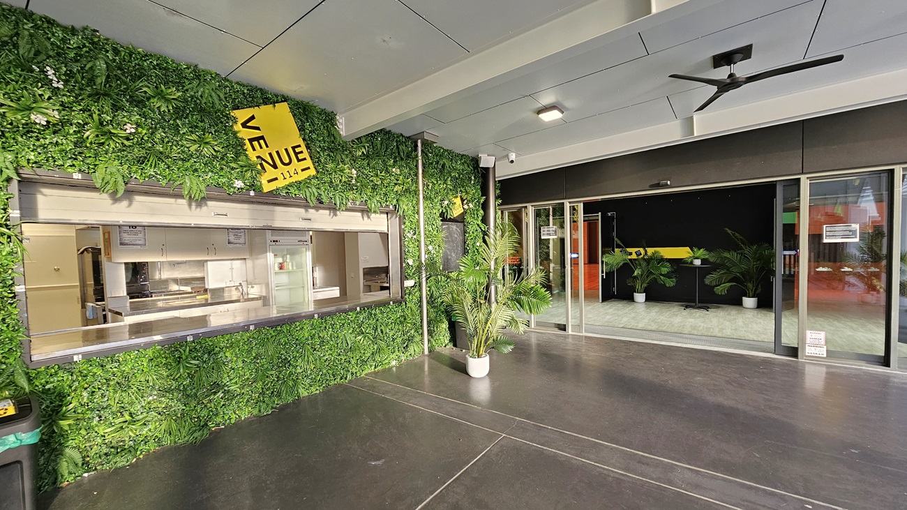 On the left, there is a service counter with a metal shutter, set within a green wall covered in artificial foliage and a yellow sign reading "Venue". On the right, there are glass sliding doors leading to an indoor area with potted plants and a black wall. In the middle, there is an open space with a dark concrete floor. The ceiling is white with recessed lights and a black ceiling fan. On the left, there is a service counter with a metal shutter, set within a green wall covered in artificial foliage and a yellow sign reading "Venue". On the right, there are glass sliding doors leading to an indoor area with potted plants and a black wall. In the middle, there is an open space with a dark concrete floor. The ceiling is white with recessed lights and a black ceiling fan.