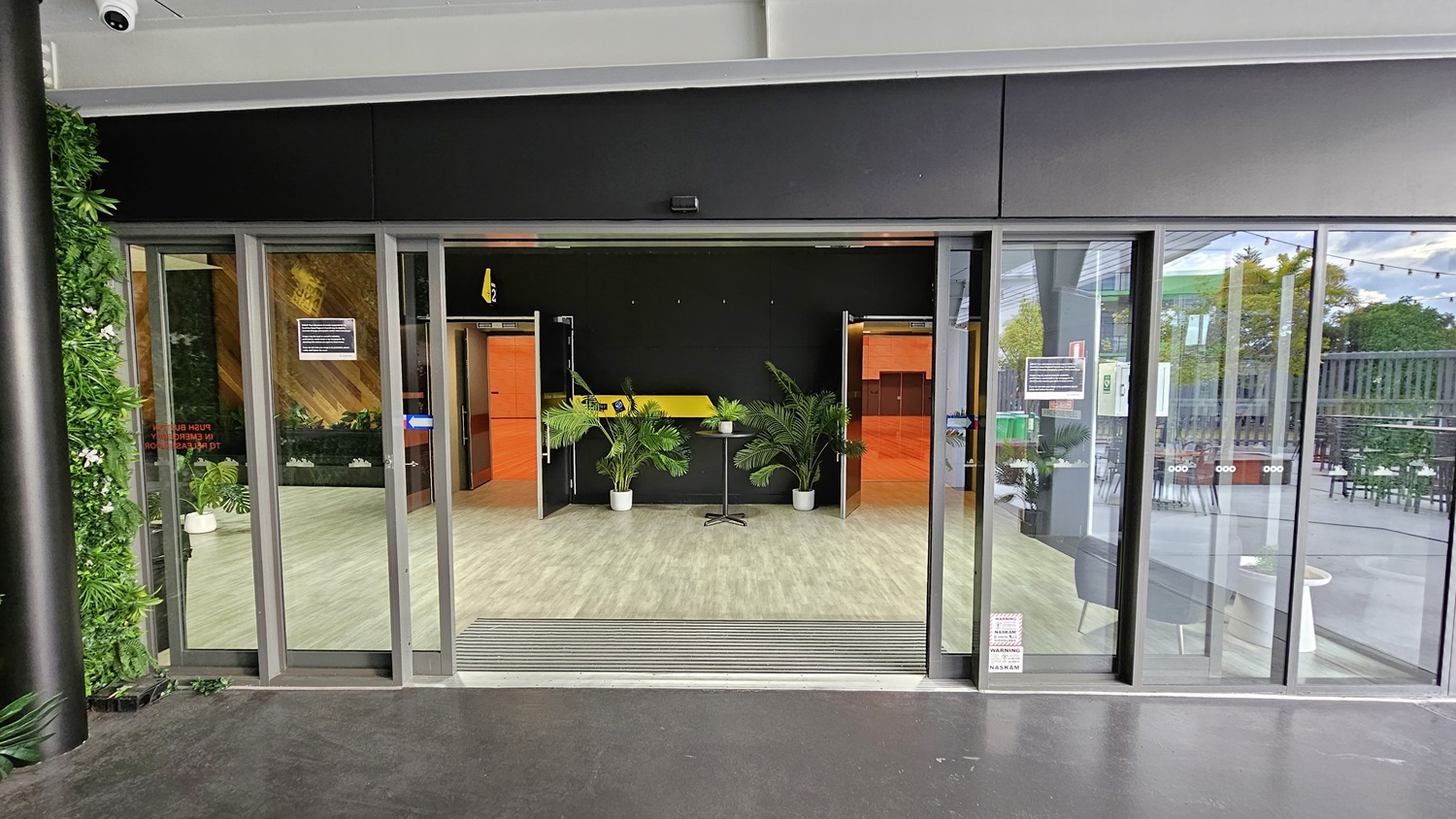 On the left, there is a green wall with artificial foliage and a black pillar. On the right, there are glass doors leading to an outdoor seating area with tables and chairs. In the middle, there is a reception area with a yellow counter, potted plants, and orange doors on either side. The floor is light grey wood-effect tile, and the ceiling is white with recessed lights. On the left, there is a green wall with artificial foliage and a black pillar. On the right, there are glass doors leading to an outdoor seating area with tables and chairs. In the middle, there is a reception area with a yellow counter, potted plants, and orange doors on either side. The floor is light grey wood-effect tile, and the ceiling is white with recessed lights.