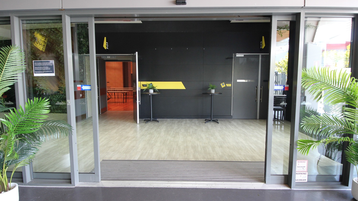 On the left, there is a green wall with artificial foliage and a small sign. On the right, there is a black wall with a door and a yellow sign above it. In the middle, there is an open entrance with glass sliding doors leading to a room with a light wood-effect tile floor. The room has a black wall with a yellow stripe, two small black tables with plants, and an open door to another room with orange walls. The ceiling is white with recessed lights. On the left, there is a green wall with artificial foliage and a small sign. On the right, there is a black wall with a door and a yellow sign above it. In the middle, there is an open entrance with glass sliding doors leading to a room with a light wood-effect tile floor. The room has a black wall with a yellow stripe, two small black tables with plants, and an open door to another room with orange walls. The ceiling is white with recessed lights.