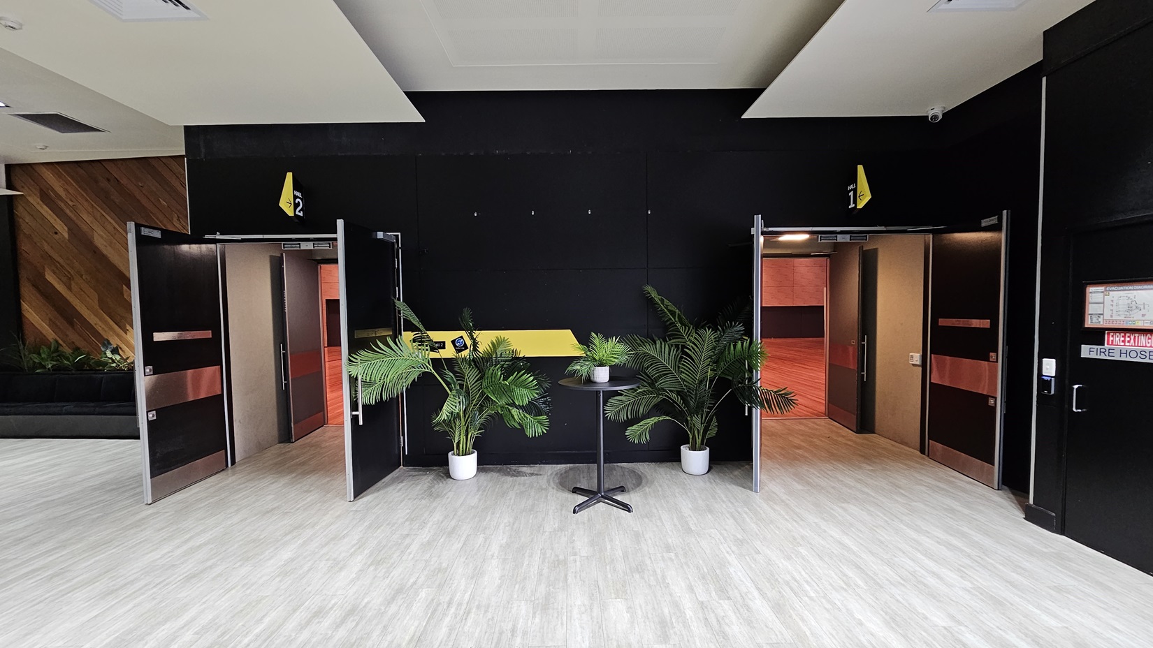 On the left, there is an open door leading to a room with red walls and wooden flooring. On the right, there is another open door leading to a similar room. In the middle, there is a black wall with a yellow stripe, two potted plants, and a small round table with a plant on it. The background features black walls with yellow signs above the doors. The floor is light grey wood, and the ceiling is white with recessed lights. On the left, there is an open door leading to a room with red walls and wooden flooring. On the right, there is another open door leading to a similar room. In the middle, there is a black wall with a yellow stripe, two potted plants, and a small round table with a plant on it. The background features black walls with yellow signs above the doors. The floor is light grey wood, and the ceiling is white with recessed lights.