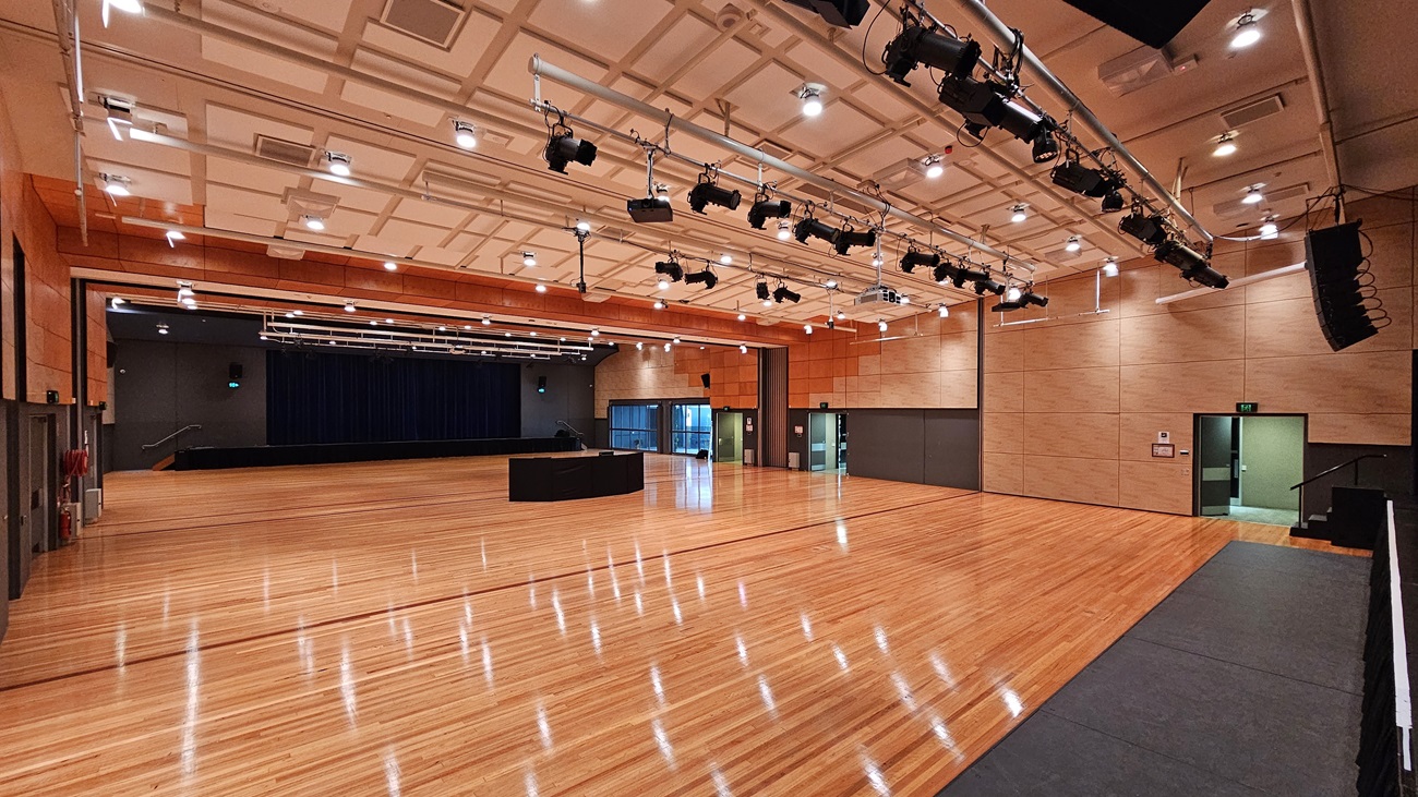 On the left, there is a stage with blue curtains and black walls, along with a fire extinguisher mounted on the wall. On the right, there is a beige wall with a green exit door. In the middle, there is a large open space with polished light wood flooring and a black equipment table. The background features beige and orange acoustic panels on the walls. The ceiling is white with a grid pattern and numerous recessed lights, along with black lighting equipment suspended from metal tracks. On the left, there is a stage with blue curtains and black walls, along with a fire extinguisher mounted on the wall. On the right, there is a beige wall with a green exit door. In the middle, there is a large open space with polished light wood flooring and a black equipment table. The background features beige and orange acoustic panels on the walls. The ceiling is white with a grid pattern and numerous recessed lights, along with black lighting equipment suspended from metal tracks.
