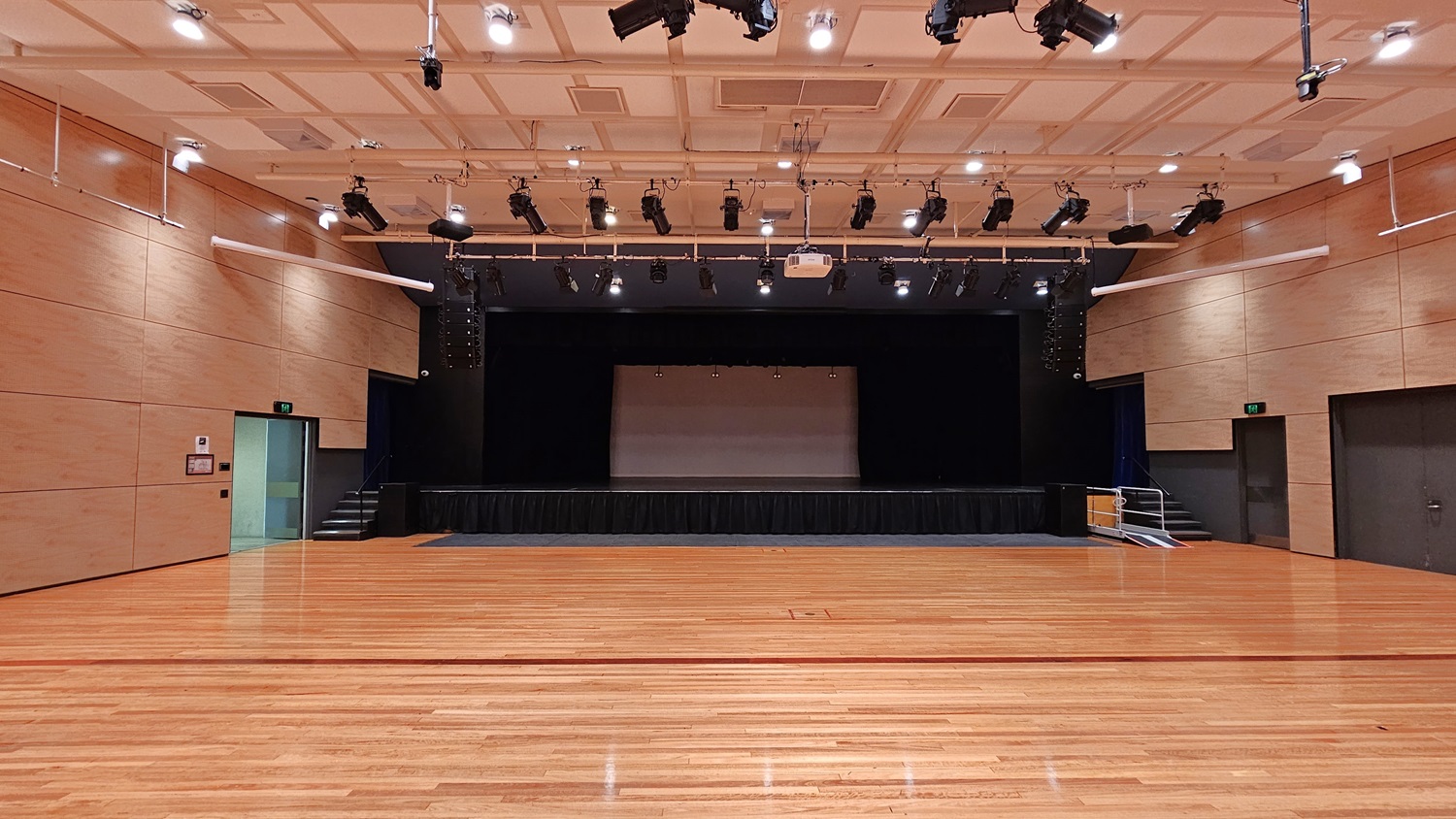 On the left, there is a beige wall with a green exit door and a fire extinguisher mounted on the wall. On the right, there is another beige wall with a dark grey door. In the middle, there is a large stage with black curtains and lighting equipment above. The background features beige walls and a black stage area. The floor is polished light wood, and the ceiling is white with a grid pattern and numerous recessed lights, along with black lighting equipment suspended from metal tracks. On the left, there is a beige wall with a green exit door and a fire extinguisher mounted on the wall. On the right, there is another beige wall with a dark grey door. In the middle, there is a large stage with black curtains and lighting equipment above. The background features beige walls and a black stage area. The floor is polished light wood, and the ceiling is white with a grid pattern and numerous recessed lights, along with black lighting equipment suspended from metal tracks.