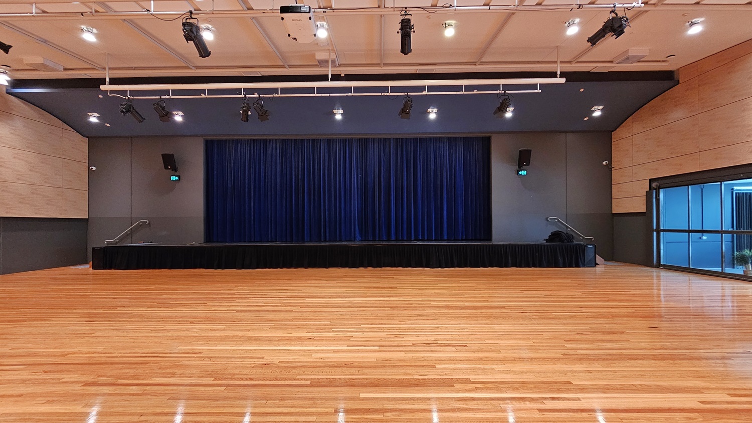 On the left is a beige wall with a black lower section and a green exit sign above a door. On the right, there is a similar beige wall with a black lower section and a green exit sign above another door. In the middle, there is a stage with blue curtains and black skirting. The background features the stage area with blue curtains. The floor is polished light wood, and the ceiling is white with a grid pattern and recessed lights, along with black lighting equipment suspended from metal tracks. On the left is a beige wall with a black lower section and a green exit sign above a door. On the right, there is a similar beige wall with a black lower section and a green exit sign above another door. In the middle, there is a stage with blue curtains and black skirting. The background features the stage area with blue curtains. The floor is polished light wood, and the ceiling is white with a grid pattern and recessed lights, along with black lighting equipment suspended from metal tracks.