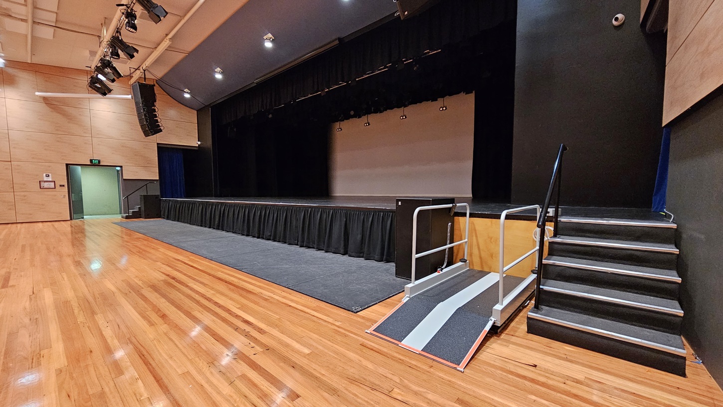 On the left, there is a beige wall with a green exit door and a fire alarm. On the right, there is a black wall with a set of stairs leading up to the stage. In the middle, there is a large stage with black curtains and a grey ramp with handrails. The background features the stage area with black curtains and lighting equipment above. The floor is polished light wood, and the ceiling is white with recessed lights and black speakers. On the left, there is a beige wall with a green exit door and a fire alarm. On the right, there is a black wall with a set of stairs leading up to the stage. In the middle, there is a large stage with black curtains and a grey ramp with handrails. The background features the stage area with black curtains and lighting equipment above. The floor is polished light wood, and the ceiling is white with recessed lights and black speakers.