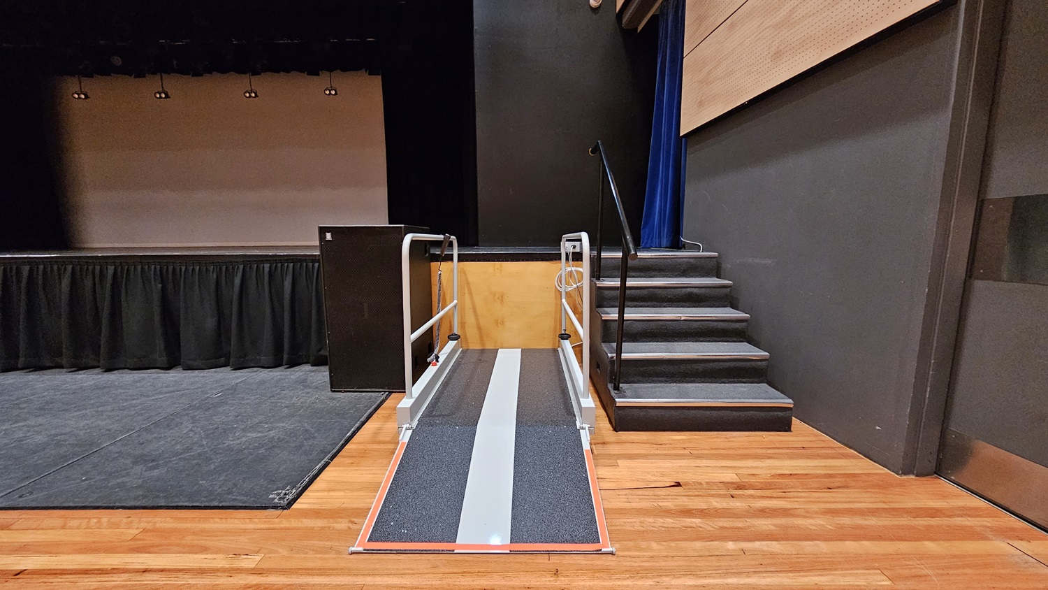 On the left, there is a stage with black curtains and a black floor. On the right, there is a set of black stairs with a handrail leading up to the stage. In the middle, there is a grey ramp with handrails on both sides, leading up to the stage. The background features a beige wall with black curtains above. The floor is polished light wood, and the ceiling is black with recessed lights. On the left, there is a stage with black curtains and a black floor. On the right, there is a set of black stairs with a handrail leading up to the stage. In the middle, there is a grey ramp with handrails on both sides, leading up to the stage. The background features a beige wall with black curtains above. The floor is polished light wood, and the ceiling is black with recessed lights.