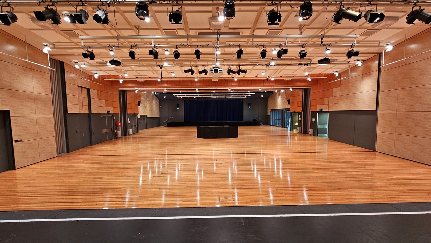 On the left, there are beige wooden walls with black doors and a fire extinguisher mounted on the wall. On the right, there are similar beige wooden walls with black doors and a green exit sign above one door. In the middle, there is a large open space with polished light wood flooring and a black stage with blue curtains in the background. The ceiling is white with a grid pattern, featuring numerous recessed lights and black stage lighting equipment. On the left, there are beige wooden walls with black doors and a fire extinguisher mounted on the wall. On the right, there are similar beige wooden walls with black doors and a green exit sign above one door. In the middle, there is a large open space with polished light wood flooring and a black stage with blue curtains in the background. The ceiling is white with a grid pattern, featuring numerous recessed lights and black stage lighting equipment.