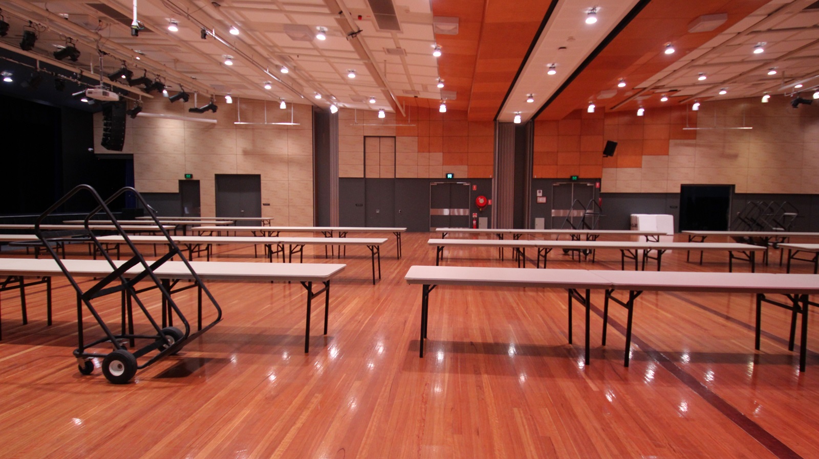 On the left, there are black double doors and a stage area with black curtains and lighting equipment. On the right, there are beige walls with black doors and a green exit sign above one door. In the middle, there are several long tables arranged in rows, with a black trolley holding folded tables. The background features beige and orange acoustic panels on the walls. The floor is polished light wood, and the ceiling is white with a grid pattern and numerous recessed lights. On the left, there are black double doors and a stage area with black curtains and lighting equipment. On the right, there are beige walls with black doors and a green exit sign above one door. In the middle, there are several long tables arranged in rows, with a black trolley holding folded tables. The background features beige and orange acoustic panels on the walls. The floor is polished light wood, and the ceiling is white with a grid pattern and numerous recessed lights.