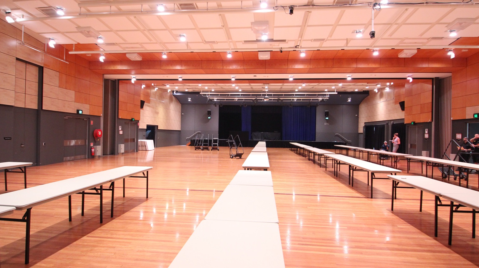 On the left, there are beige wooden walls with black double doors and a fire extinguisher mounted on the wall. On the right, there are similar beige wooden walls with black doors and a green exit sign above one door. In the middle, there are long white tables arranged in rows on a polished light wood floor. The background features a stage with blue curtains and black skirting. The ceiling is white with a grid pattern, recessed lights, and black stage lighting equipment. On the left, there are beige wooden walls with black double doors and a fire extinguisher mounted on the wall. On the right, there are similar beige wooden walls with black doors and a green exit sign above one door. In the middle, there are long white tables arranged in rows on a polished light wood floor. The background features a stage with blue curtains and black skirting. The ceiling is white with a grid pattern, recessed lights, and black stage lighting equipment.