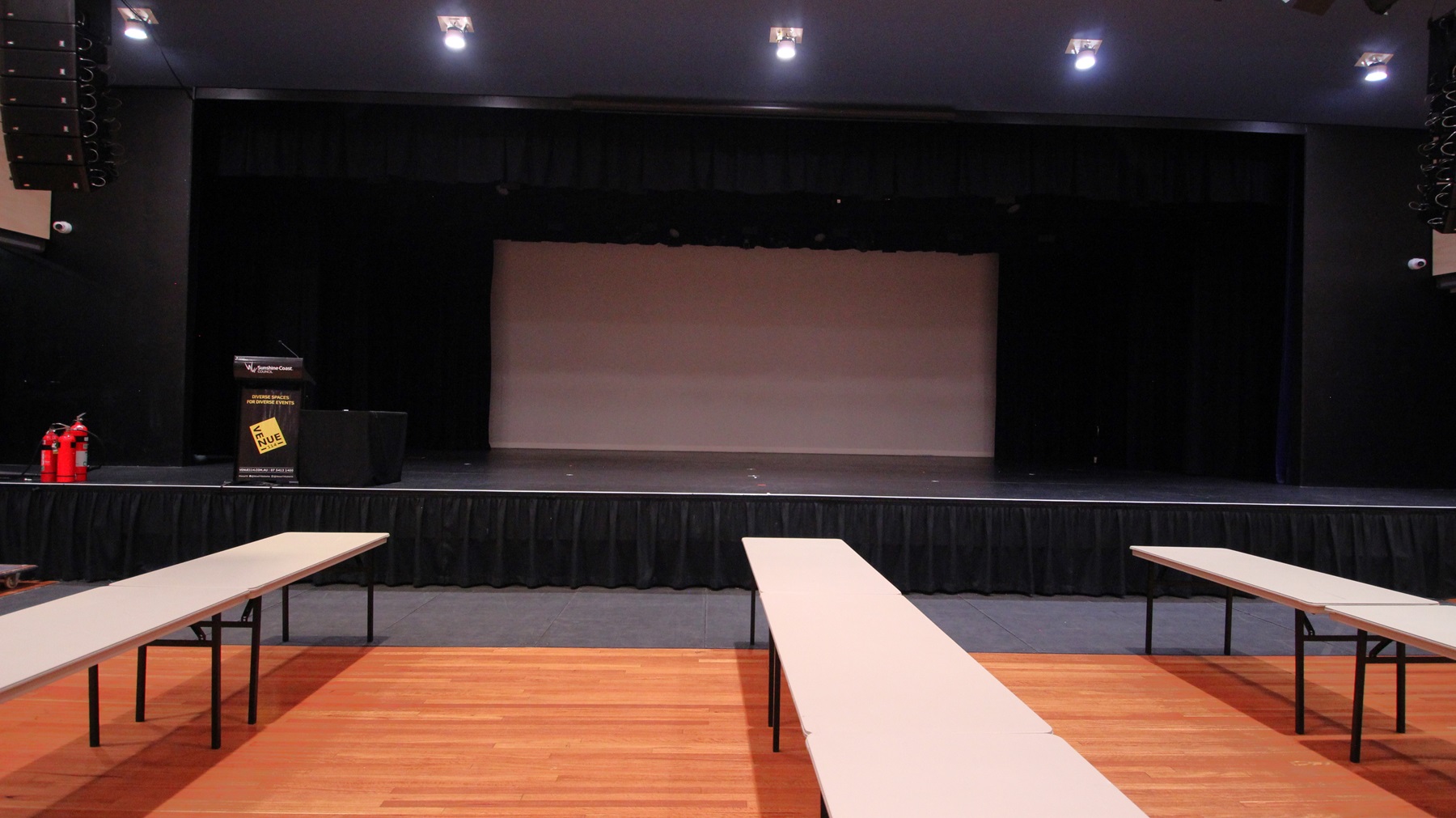 On the left, there are two red fire extinguishers on the floor next to a black podium with a sign. On the right, there is a black wall with stage lighting equipment. In the middle, there is a large stage with black curtains and a grey backdrop. The foreground features long white tables arranged in rows on a polished light wood floor. The ceiling is dark with recessed lights and additional stage lighting equipment. On the left, there are two red fire extinguishers on the floor next to a black podium with a sign. On the right, there is a black wall with stage lighting equipment. In the middle, there is a large stage with black curtains and a grey backdrop. The foreground features long white tables arranged in rows on a polished light wood floor. The ceiling is dark with recessed lights and additional stage lighting equipment.