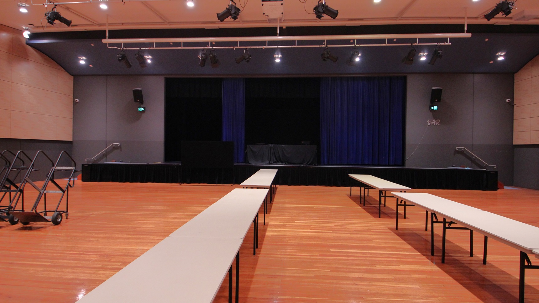 On the left, there are beige walls with black lower sections and two trolleys holding folded tables. On the right, there are similar beige walls with black lower sections and a green exit sign above a door. In the middle, there are long white tables arranged in rows on a polished light wood floor. The background features a stage with blue curtains and black skirting. The ceiling is white with recessed lights and black stage lighting equipment. On the left, there are beige walls with black lower sections and two trolleys holding folded tables. On the right, there are similar beige walls with black lower sections and a green exit sign above a door. In the middle, there are long white tables arranged in rows on a polished light wood floor. The background features a stage with blue curtains and black skirting. The ceiling is white with recessed lights and black stage lighting equipment.