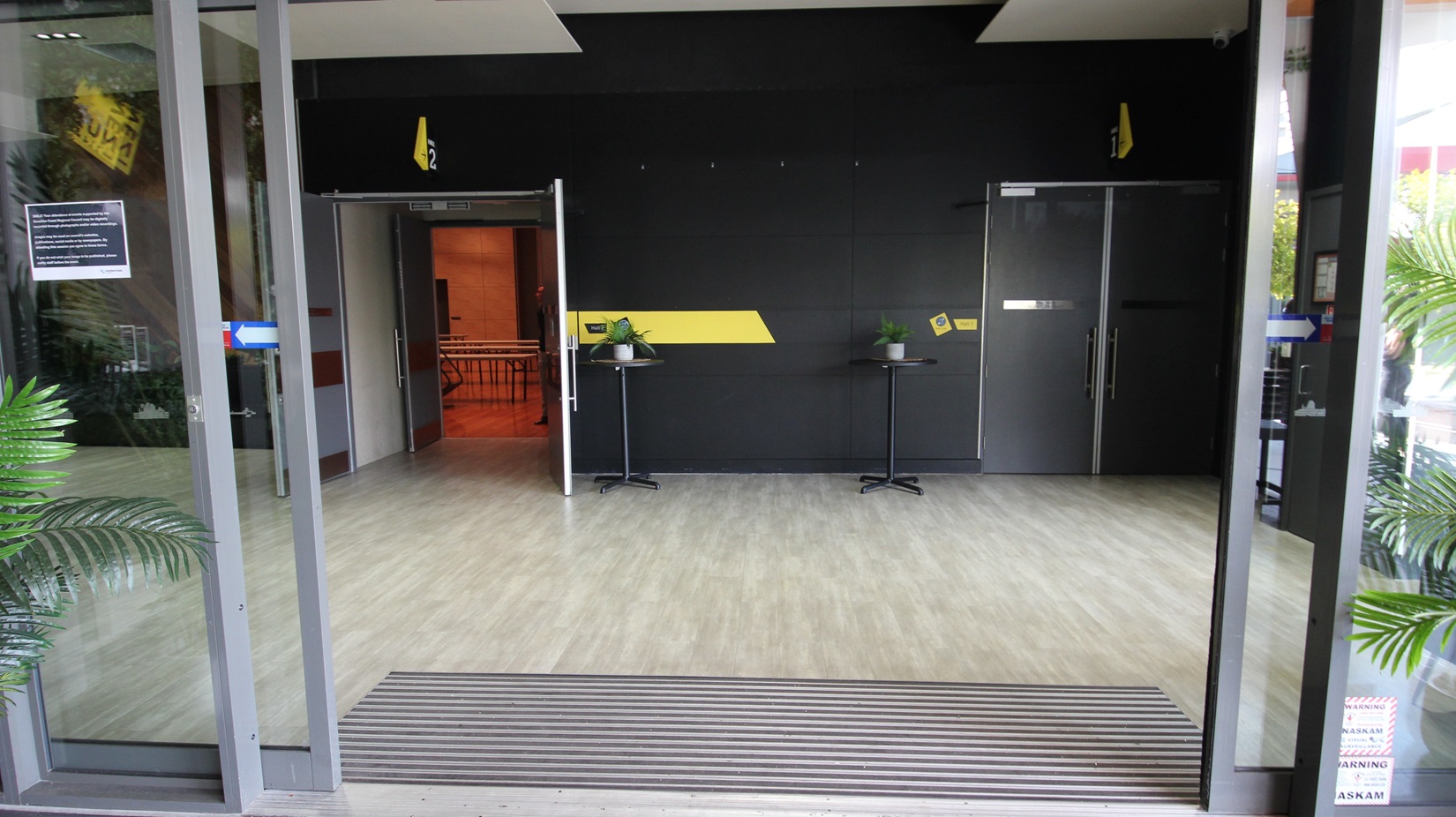 On the left, there is a glass door with a potted plant next to it. On the right, there is another glass door with a potted plant beside it. In the middle, there is a black wall with a yellow diagonal stripe, two small round tables with plants on them, and two grey doors. The floor is light wood, and the ceiling is black with recessed lights. On the left, there is a glass door with a potted plant next to it. On the right, there is another glass door with a potted plant beside it. In the middle, there is a black wall with a yellow diagonal stripe, two small round tables with plants on them, and two grey doors. The floor is light wood, and the ceiling is black with recessed lights.
