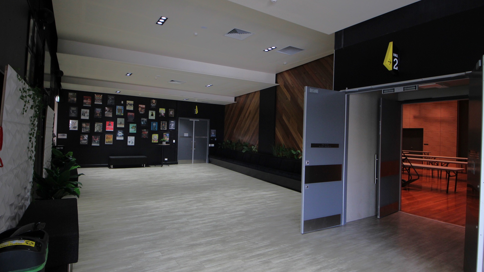 On the left, there is a black bench with potted plants and a wall covered with framed pictures. On the right, there are two open grey doors leading to a room with wooden flooring and tables. In the middle, there is an open space with light wood flooring. The background features a black wall with a yellow sign and a section of wooden panelling. The ceiling is white with recessed lights and ventilation panels. On the left, there is a black bench with potted plants and a wall covered with framed pictures. On the right, there are two open grey doors leading to a room with wooden flooring and tables. In the middle, there is an open space with light wood flooring. The background features a black wall with a yellow sign and a section of wooden panelling. The ceiling is white with recessed lights and ventilation panels.