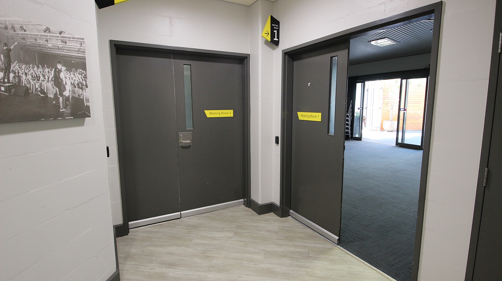 On the left, there is a black and white photograph of a crowd on a light grey wall. Below the photograph, there are two grey double doors with yellow signs above them. On the right, there is an open doorway leading to a room with dark carpet and large windows. The floor is light grey wood, and the ceiling is grey with recessed lights and ventilation panels. On the left, there is a black and white photograph of a crowd on a light grey wall. Below the photograph, there are two grey double doors with yellow signs above them. On the right, there is an open doorway leading to a room with dark carpet and large windows. The floor is light grey wood, and the ceiling is grey with recessed lights and ventilation panels.