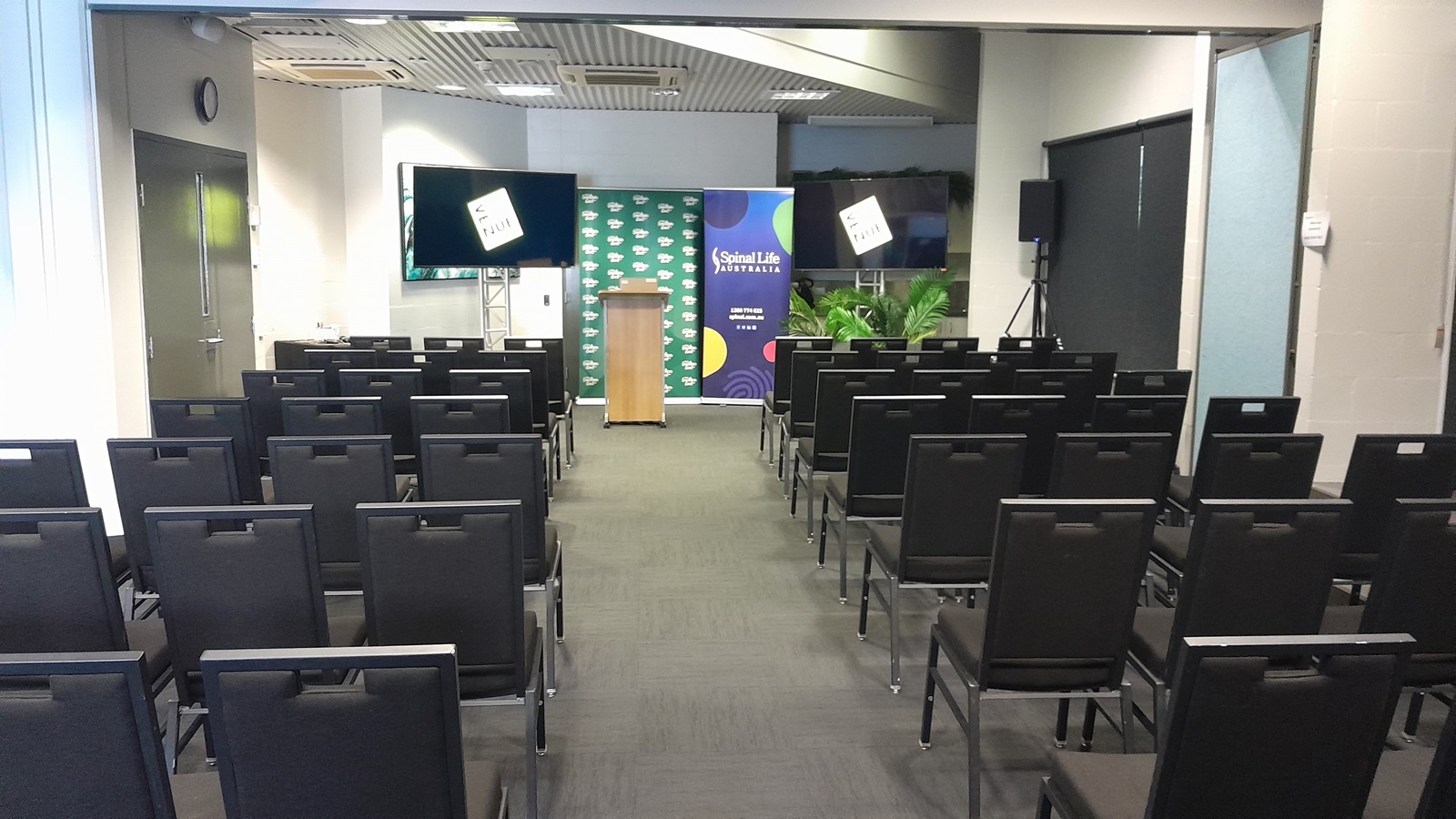 On the left, there is a wall with a closed door and a mounted television screen. On the right, there is a wall with a window covered by a black roller blind and a potted plant in front of it. In the middle, there are rows of black chairs facing a wooden podium with a microphone, flanked by two banners and another television screen. The floor is covered in dark grey carpet, and the ceiling is white with recessed lights and ventilation panels. On the left, there is a wall with a closed door and a mounted television screen. On the right, there is a wall with a window covered by a black roller blind and a potted plant in front of it. In the middle, there are rows of black chairs facing a wooden podium with a microphone, flanked by two banners and another television screen. The floor is covered in dark grey carpet, and the ceiling is white with recessed lights and ventilation panels.