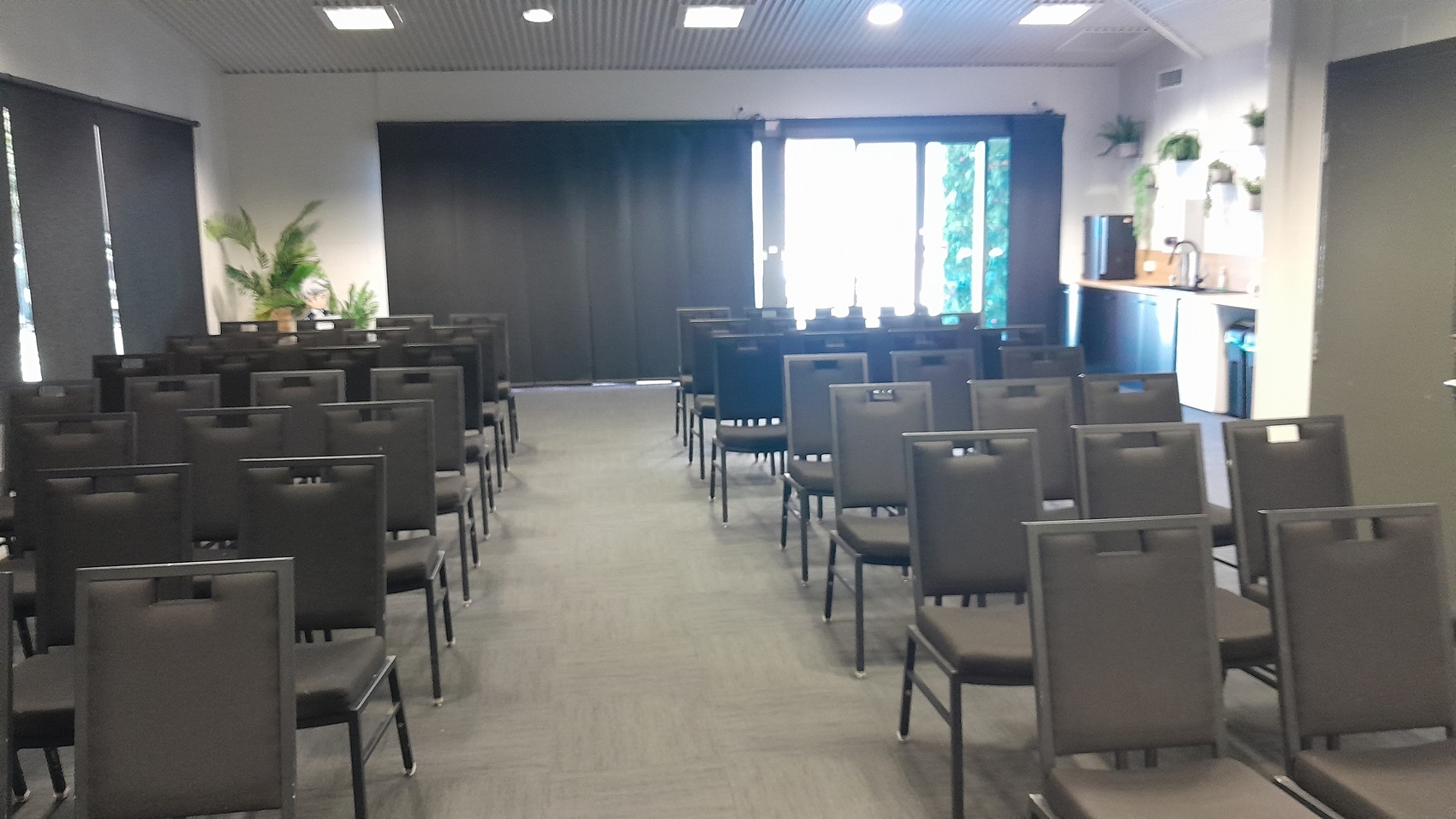 On the left, there are rows of black chairs facing forward, with a wall featuring black roller blinds and potted plants. On the right, there are more rows of black chairs, a countertop with a coffee machine, cups, and potted plants above. In the background, there is a large window with black curtains partially open, revealing greenery outside. The floor is covered in dark grey carpet, and the ceiling is white with recessed lights and ventilation panels. On the left, there are rows of black chairs facing forward, with a wall featuring black roller blinds and potted plants. On the right, there are more rows of black chairs, a countertop with a coffee machine, cups, and potted plants above. In the background, there is a large window with black curtains partially open, revealing greenery outside. The floor is covered in dark grey carpet, and the ceiling is white with recessed lights and ventilation panels.