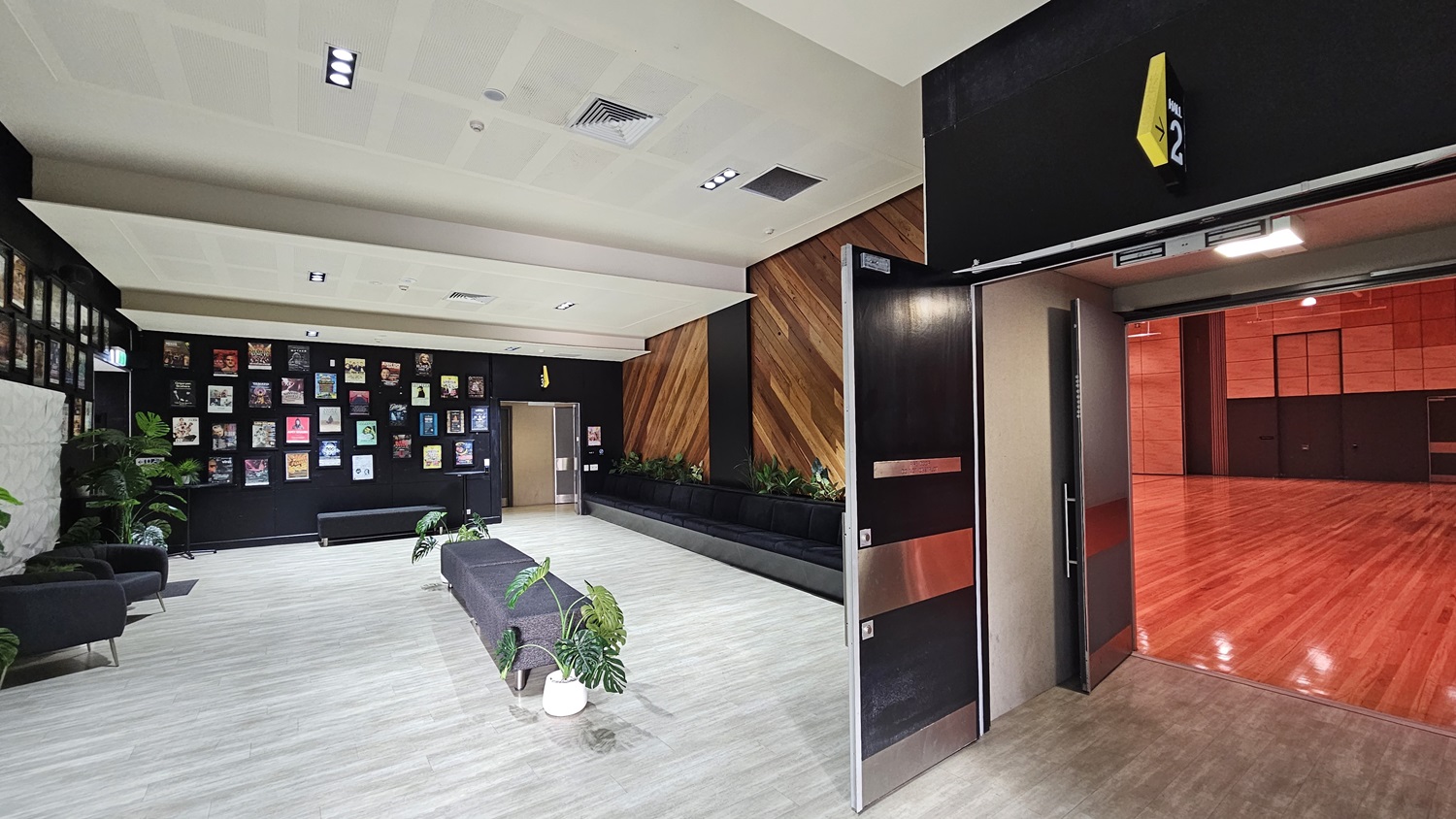 On the left, there is a black bench with potted plants and a wall covered with framed pictures. On the right, there are two open grey doors leading to a room with a wooden floor and red walls. In the middle, there are two black armchairs with a small table and potted plants. The background features a black wall with diagonal wooden panels and additional potted plants. The floor is light grey wood, and the ceiling is white with recessed lights and ventilation panels. On the left, there is a black bench with potted plants and a wall covered with framed pictures. On the right, there are two open grey doors leading to a room with a wooden floor and red walls. In the middle, there are two black armchairs with a small table and potted plants. The background features a black wall with diagonal wooden panels and additional potted plants. The floor is light grey wood, and the ceiling is white with recessed lights and ventilation panels.
