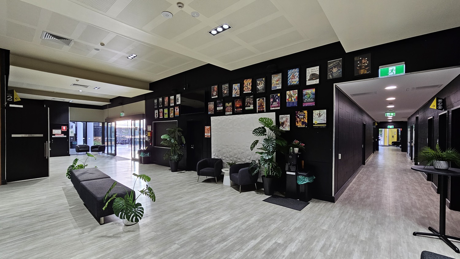 On the left, there is a long black bench with potted plants on the floor and a wall with framed posters above. On the right, there is a corridor with a light grey floor leading to a brightly lit area, and a black wall with a whiteboard and two black armchairs with potted plants. In the background, there are glass doors opening to an outdoor area with more seating. The floor is light grey wood, and the ceiling is white with recessed lights and ventilation panels. On the left, there is a long black bench with potted plants on the floor and a wall with framed posters above. On the right, there is a corridor with a light grey floor leading to a brightly lit area, and a black wall with a whiteboard and two black armchairs with potted plants. In the background, there are glass doors opening to an outdoor area with more seating. The floor is light grey wood, and the ceiling is white with recessed lights and ventilation panels.