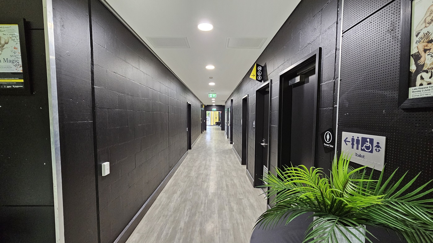 On the left, there is a black wall with a framed poster near the top. On the right, there is a black wall with a door marked with a toilet sign and a potted plant in the foreground. In the middle, there is a long corridor with a light grey wood floor leading to a distant area with a green exit sign above the door. The ceiling is white with recessed lights and ventilation panels. On the left, there is a black wall with a framed poster near the top. On the right, there is a black wall with a door marked with a toilet sign and a potted plant in the foreground. In the middle, there is a long corridor with a light grey wood floor leading to a distant area with a green exit sign above the door. The ceiling is white with recessed lights and ventilation panels.