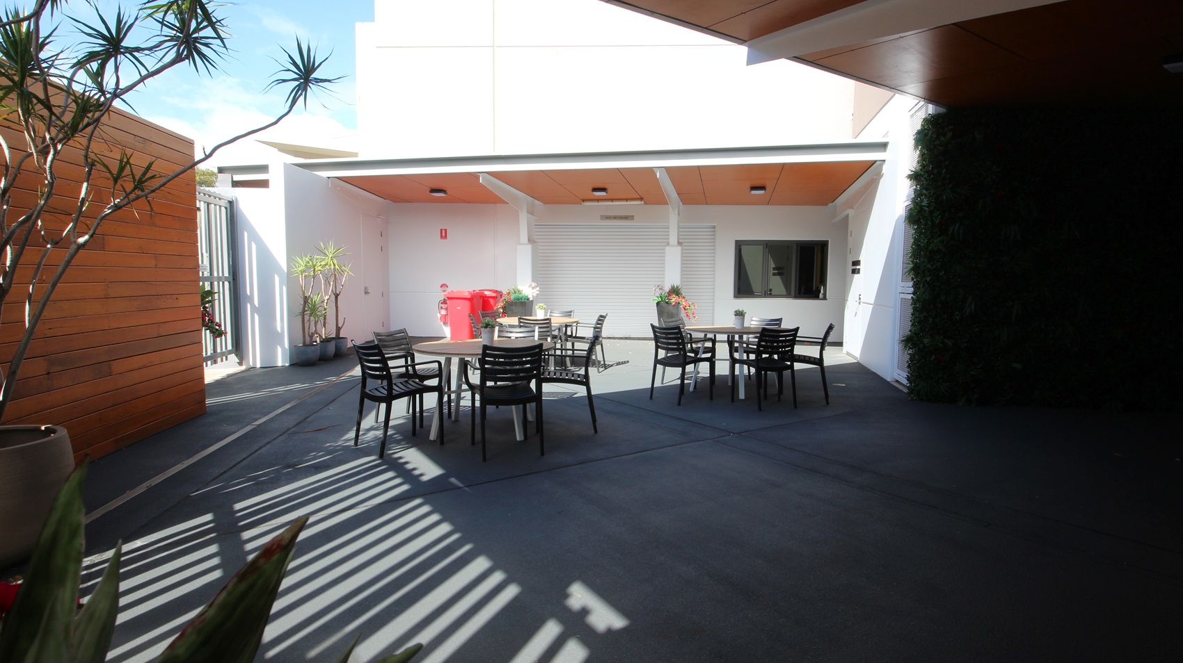 On the left, there is a wooden wall with a tall potted plant in front of it. On the right, there is a dark wall covered with greenery. In the middle, there are several black tables with black chairs arranged around them. In the background, there is a white building with a wooden ceiling overhang, a red fire extinguisher, and potted plants near a closed white roller door. The floor is dark grey concrete, and the ceiling under the overhang is wooden with recessed lights. On the left, there is a wooden wall with a tall potted plant in front of it. On the right, there is a dark wall covered with greenery. In the middle, there are several black tables with black chairs arranged around them. In the background, there is a white building with a wooden ceiling overhang, a red fire extinguisher, and potted plants near a closed white roller door. The floor is dark grey concrete, and the ceiling under the overhang is wooden with recessed lights.