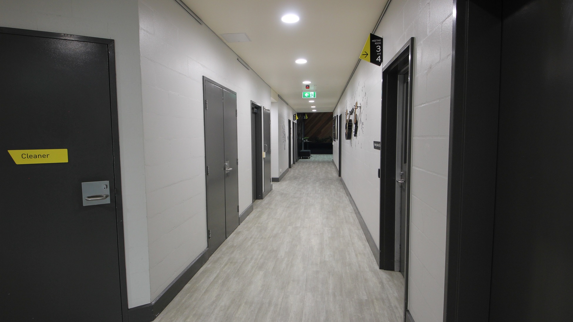 On the left, there are grey doors with yellow signs, including one marked "Cleaner." On the right, there are more grey doors, one with a sign and hooks holding cleaning equipment. In the middle, a long corridor with a light grey wood-effect floor leads to a distant area with a green exit sign above a door. The ceiling is white with recessed lights and ventilation panels. On the left, there are grey doors with yellow signs, including one marked "Cleaner." On the right, there are more grey doors, one with a sign and hooks holding cleaning equipment. In the middle, a long corridor with a light grey wood-effect floor leads to a distant area with a green exit sign above a door. The ceiling is white with recessed lights and ventilation panels.