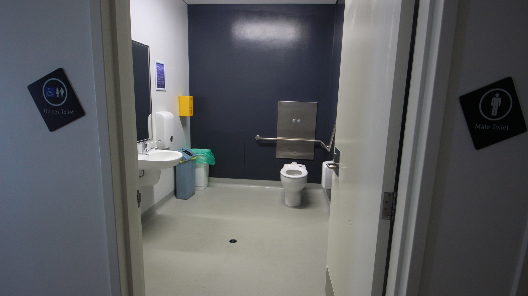 On the left, there is a white sink with a soap dispenser above it, a paper towel dispenser, and a green bin below. On the right, there is a white wall with a handrail extending from the back wall. In the middle, there is a white toilet with a metal flush panel above it and a yellow bin to the left. The floor is light grey, and the ceiling is white with no visible fixtures. The walls are a mix of white and dark blue. On the left, there is a white sink with a soap dispenser above it, a paper towel dispenser, and a green bin below. On the right, there is a white wall with a handrail extending from the back wall. In the middle, there is a white toilet with a metal flush panel above it and a yellow bin to the left. The floor is light grey, and the ceiling is white with no visible fixtures. The walls are a mix of white and dark blue.