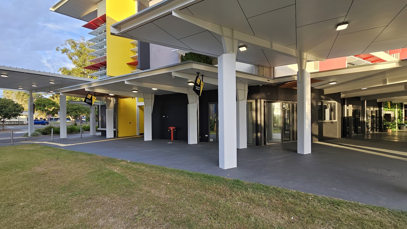 On the left, there is a yellow wall with a door and a black wall with a yellow sign. On the right, there are glass revolving doors leading into a building with a black wall. In the middle, there are white pillars supporting a white ceiling with recessed lights. The floor is grey concrete, and the foreground has a grassy area. The background includes additional white pillars and a yellow vertical structure with balconies. On the left, there is a yellow wall with a door and a black wall with a yellow sign. On the right, there are glass revolving doors leading into a building with a black wall. In the middle, there are white pillars supporting a white ceiling with recessed lights. The floor is grey concrete, and the foreground has a grassy area. The background includes additional white pillars and a yellow vertical structure with balconies.