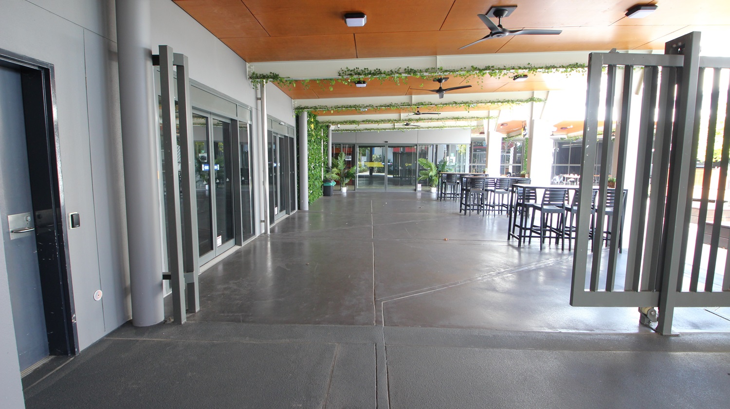 On the left, there are grey walls with glass doors and windows, and a black door. On the right, there are tall metal gates and a seating area with high tables and chairs. In the middle, there is a wide walkway with a dark concrete floor leading to more glass doors and potted plants. The ceiling is wooden with recessed lights and ceiling fans, and it has green vines hanging from it. On the left, there are grey walls with glass doors and windows, and a black door. On the right, there are tall metal gates and a seating area with high tables and chairs. In the middle, there is a wide walkway with a dark concrete floor leading to more glass doors and potted plants. The ceiling is wooden with recessed lights and ceiling fans, and it has green vines hanging from it.