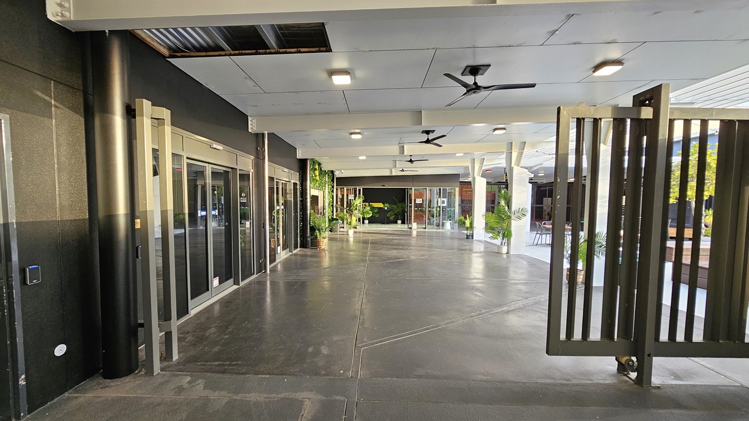 On the left, there are black walls with large glass windows and doors, and a black pillar. On the right, there are tall metal gates and a seating area with tables, chairs, and potted plants. In the middle, there is a wide walkway with a dark concrete floor leading to more glass doors and greenery. The ceiling is white with recessed lights and ceiling fans. On the left, there are black walls with large glass windows and doors, and a black pillar. On the right, there are tall metal gates and a seating area with tables, chairs, and potted plants. In the middle, there is a wide walkway with a dark concrete floor leading to more glass doors and greenery. The ceiling is white with recessed lights and ceiling fans.