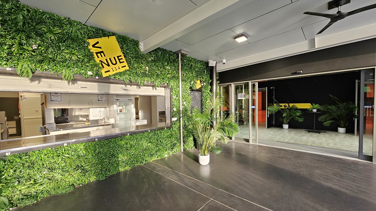 On the left, there is a green wall covered with artificial foliage and a yellow sign that reads "VENUE." Below the sign, there is a serving window with a kitchen visible inside. On the right, there are glass sliding doors leading to an indoor area with a black and yellow wall, potted plants, and a light-coloured floor. In the middle, there is a dark concrete floor. The ceiling is white with recessed lights and a ceiling fan. On the left, there is a green wall covered with artificial foliage and a yellow sign that reads "VENUE." Below the sign, there is a serving window with a kitchen visible inside. On the right, there are glass sliding doors leading to an indoor area with a black and yellow wall, potted plants, and a light-coloured floor. In the middle, there is a dark concrete floor. The ceiling is white with recessed lights and a ceiling fan.
