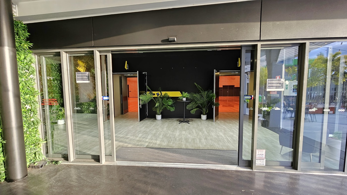 On the left, there is a green wall covered with artificial foliage. On the right, there are glass sliding doors leading to an outdoor seating area with tables and chairs. In the middle, there is an entrance with glass sliding doors opening into a room with a light wood-effect tile floor, black walls, and potted plants. The ceiling inside is white with recessed lights. On the left, there is a green wall covered with artificial foliage. On the right, there are glass sliding doors leading to an outdoor seating area with tables and chairs. In the middle, there is an entrance with glass sliding doors opening into a room with a light wood-effect tile floor, black walls, and potted plants. The ceiling inside is white with recessed lights.
