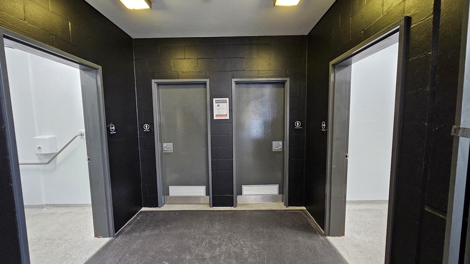 On the left, there is a grey door with a white grab bar on the adjacent wall. On the right, there is another grey door with a white wall next to it. In the middle, there are two grey doors set in a black wall, each with a small sign indicating restrooms. The floor is a mix of dark and light grey concrete, and the ceiling is white with recessed rectangular lights. On the left, there is a grey door with a white grab bar on the adjacent wall. On the right, there is another grey door with a white wall next to it. In the middle, there are two grey doors set in a black wall, each with a small sign indicating restrooms. The floor is a mix of dark and light grey concrete, and the ceiling is white with recessed rectangular lights.