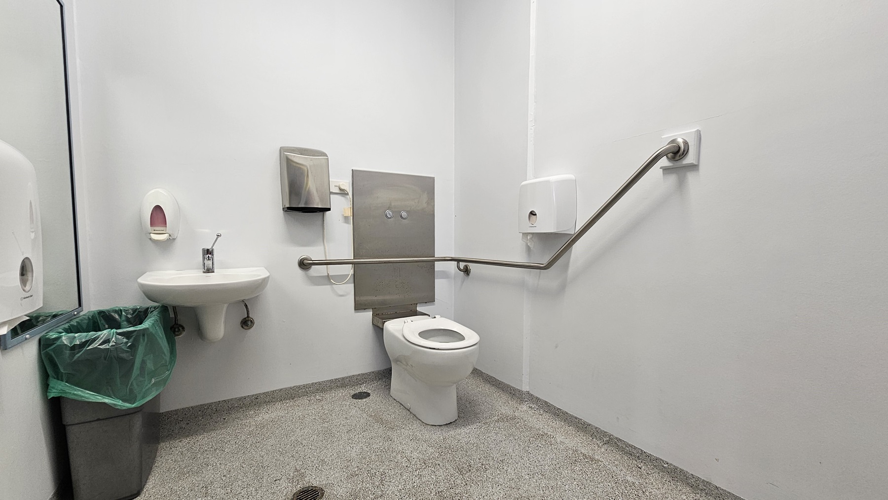 On the left, there is a white sink with a soap dispenser above it, a paper towel dispenser, and a green bin below. On the right, there is a white toilet with a metal grab bar extending along the wall. In the middle, there is a grey panel behind the toilet with two flush buttons. The floor is speckled grey, and the walls and ceiling are white. On the left, there is a white sink with a soap dispenser above it, a paper towel dispenser, and a green bin below. On the right, there is a white toilet with a metal grab bar extending along the wall. In the middle, there is a grey panel behind the toilet with two flush buttons. The floor is speckled grey, and the walls and ceiling are white.