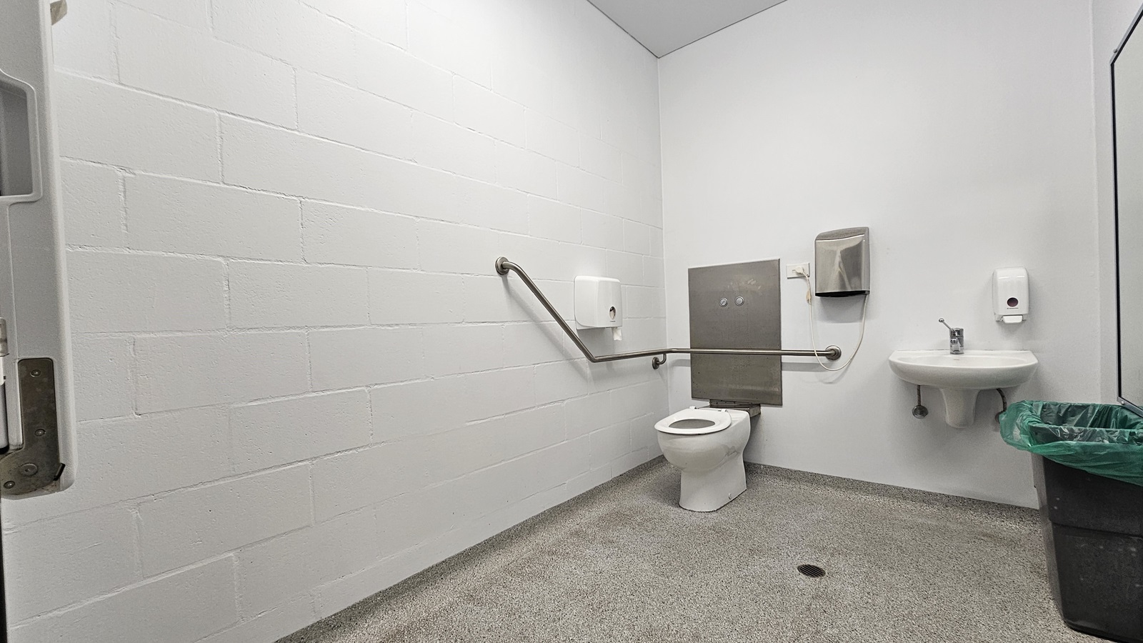On the left, there is a white wall with a metal grab bar extending horizontally. On the right, there is a white sink with a soap dispenser above it, a paper towel dispenser, and a green bin below. In the middle, there is a white toilet with a metal grab bar on the wall behind it and a grey panel with two flush buttons. The floor is speckled grey, and the ceiling is white. On the left, there is a white wall with a metal grab bar extending horizontally. On the right, there is a white sink with a soap dispenser above it, a paper towel dispenser, and a green bin below. In the middle, there is a white toilet with a metal grab bar on the wall behind it and a grey panel with two flush buttons. The floor is speckled grey, and the ceiling is white.
