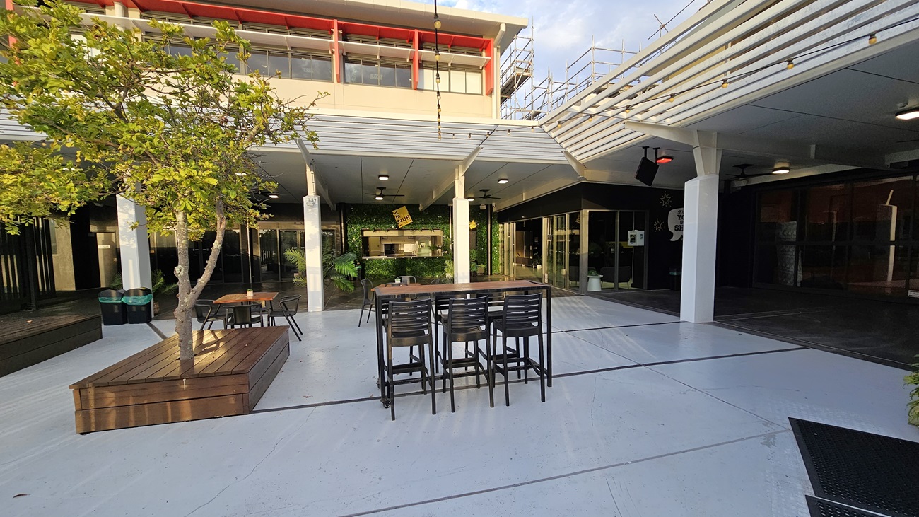 On the left, there is a wooden bench surrounding a small tree and two green bins. On the right, there is a covered area with black walls and glass doors. In the middle, there is an outdoor seating area with a tall table and several chairs. The background features a building with large windows and a red trim on the upper floor. The floor is light grey concrete, and the ceiling of the covered area is white with recessed lights. On the left, there is a wooden bench surrounding a small tree and two green bins. On the right, there is a covered area with black walls and glass doors. In the middle, there is an outdoor seating area with a tall table and several chairs. The background features a building with large windows and a red trim on the upper floor. The floor is light grey concrete, and the ceiling of the covered area is white with recessed lights.